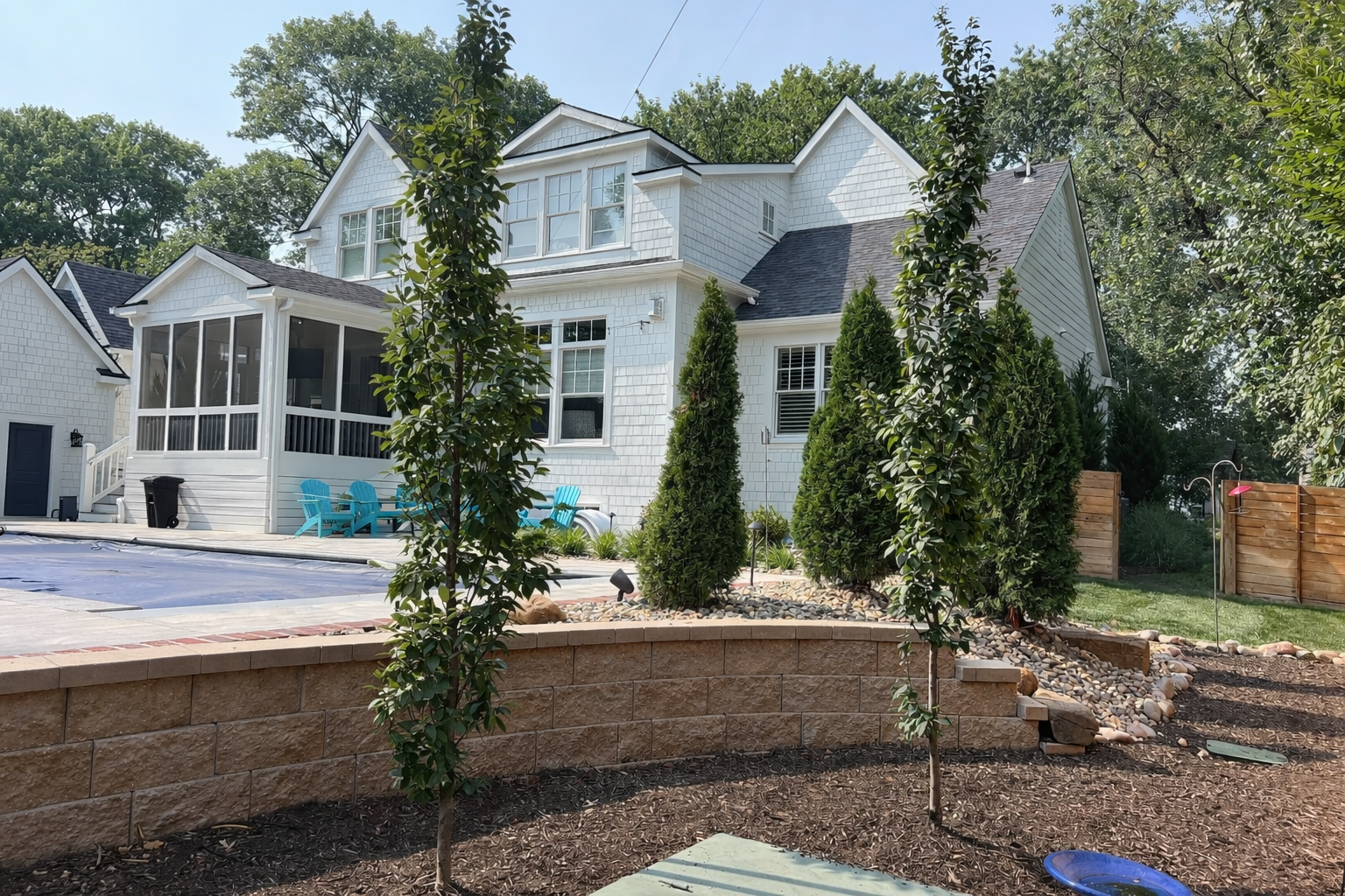 Backyard view of a white house with multiple windows, a screened porch, and a backyard landscaped with tall shrubs, trees, and a garden area with rocks and mulch.