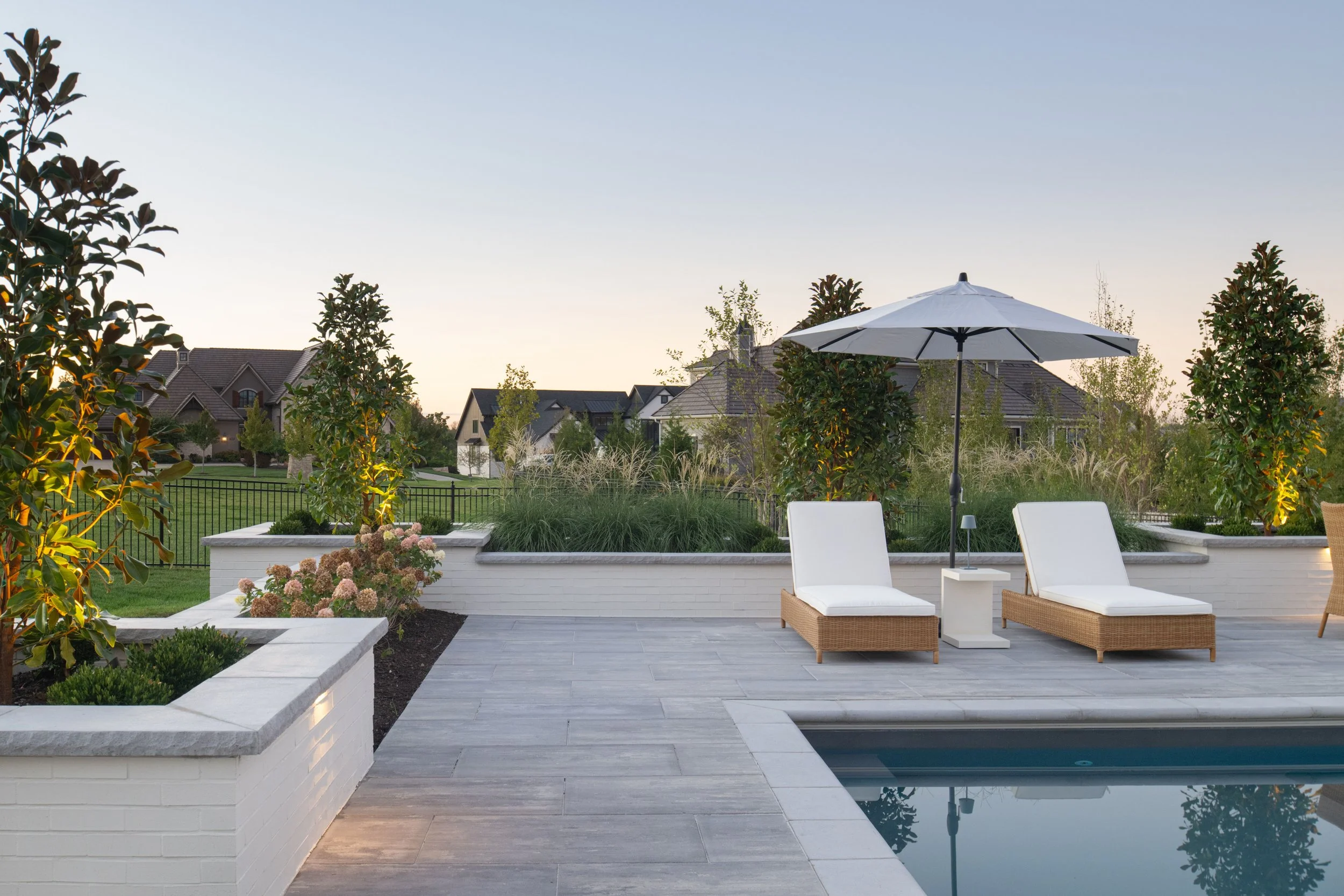Backyard patio with white chaise lounges, a white umbrella, and a small white side table near a swimming pool, surrounded by greenery and suburban houses at dusk.