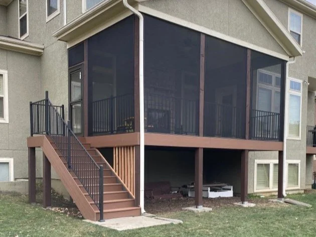 A screened porch attached to a two-story house with stairs leading up to it, enclosed with dark screen panels and a metal railing.