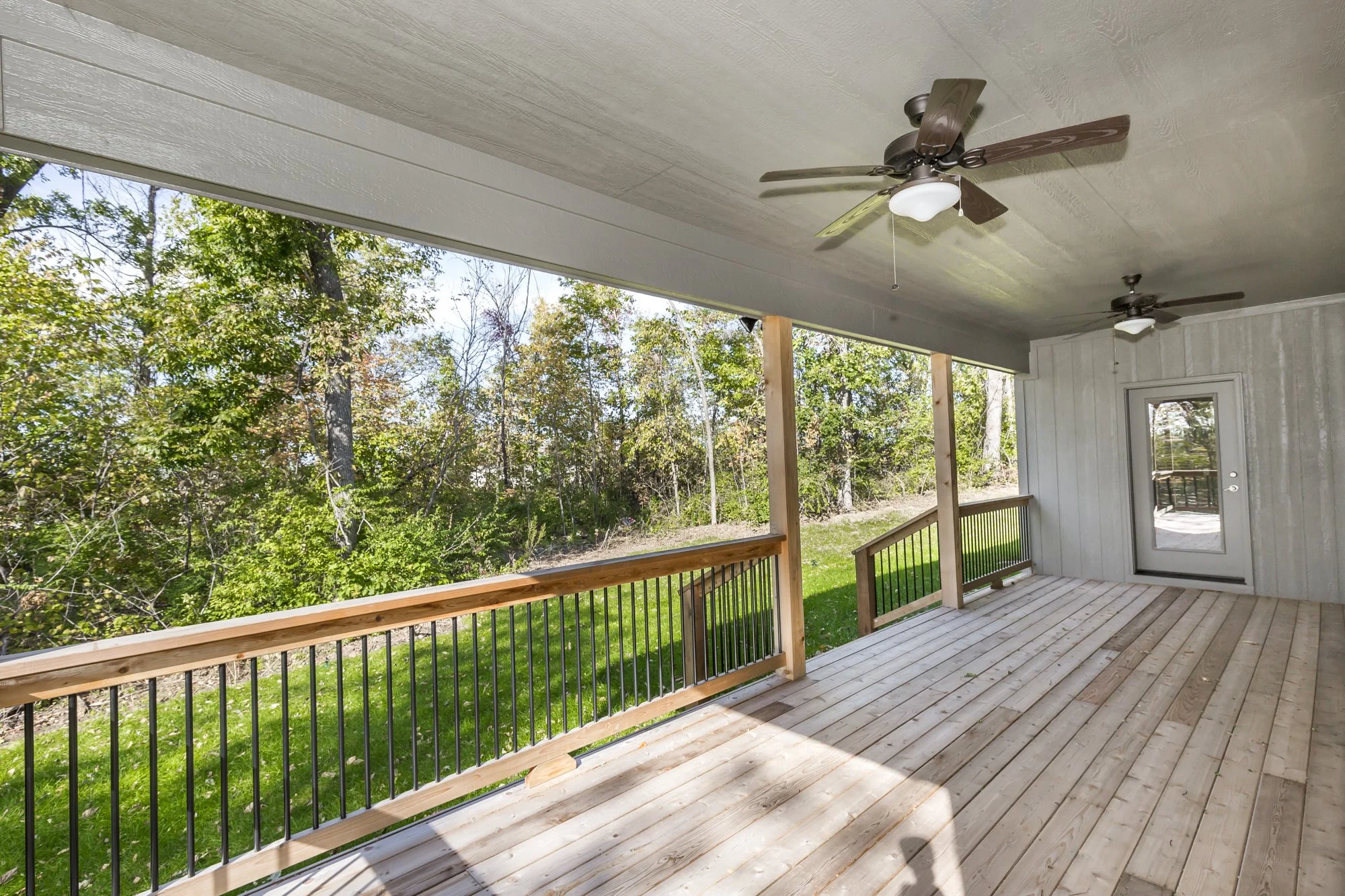 Image of a covered wooden porch with two ceiling fans, a door, and a view of trees and grass outside.