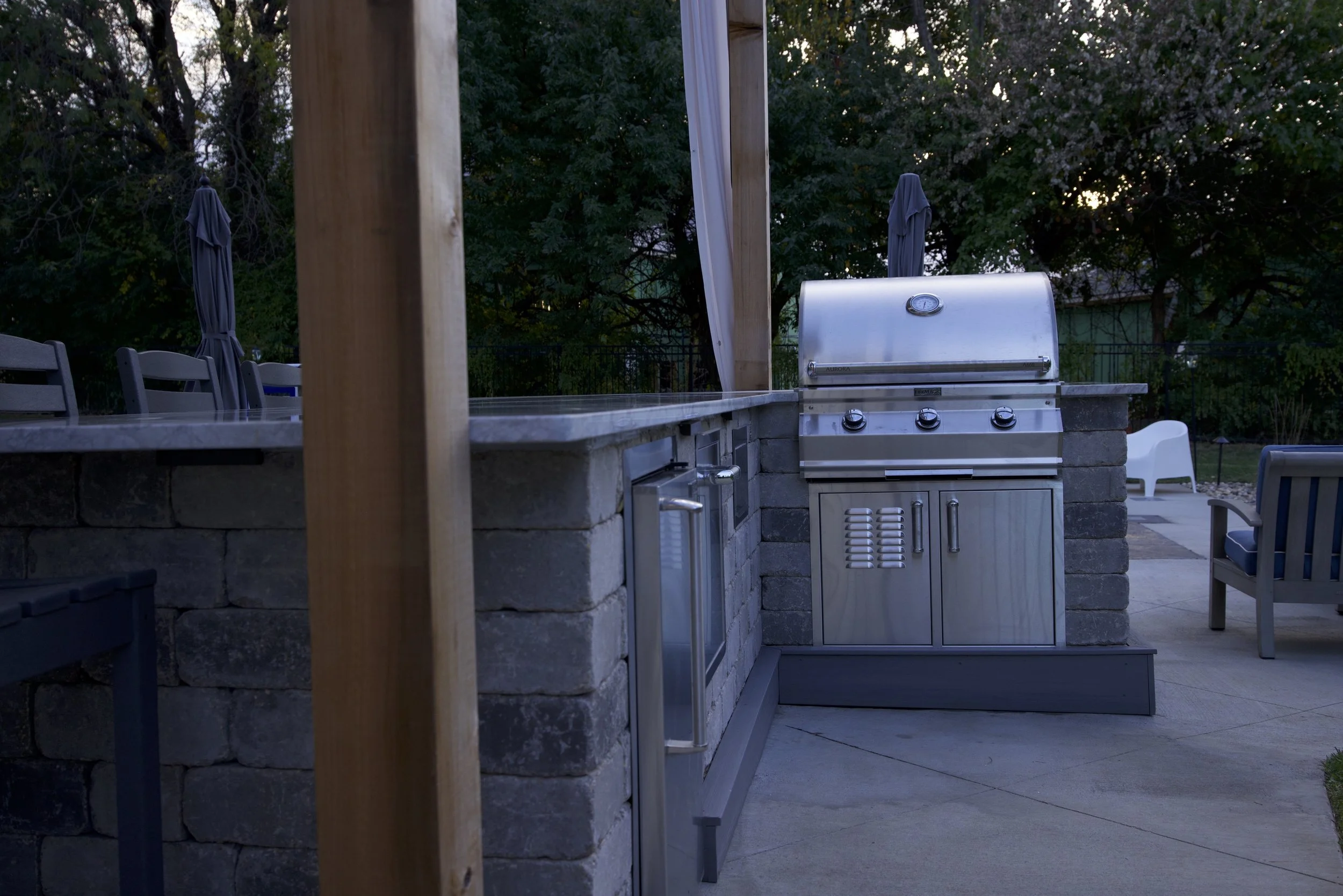 Outdoor kitchen area with a built-in stainless steel grill, surrounded by a stone counter and chairs, with trees and umbrellas in the background during dusk.