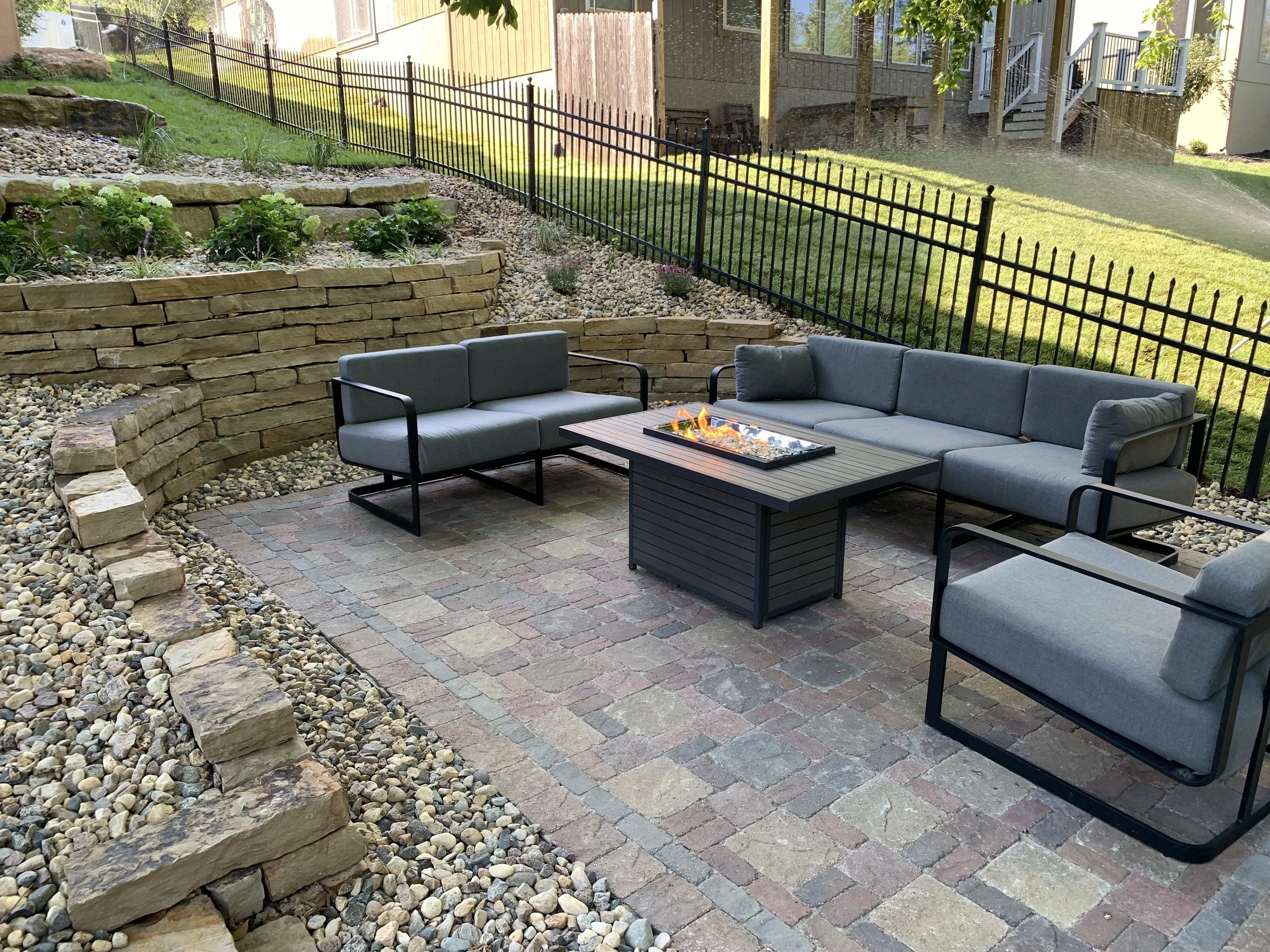 A backyard patio with three gray cushioned outdoor chairs arranged around a rectangular fire pit table on a brick and stone paved surface, with a tiered stone retaining wall and landscaped garden with green plants and gravel in the background, enclosed by a black metal fence.