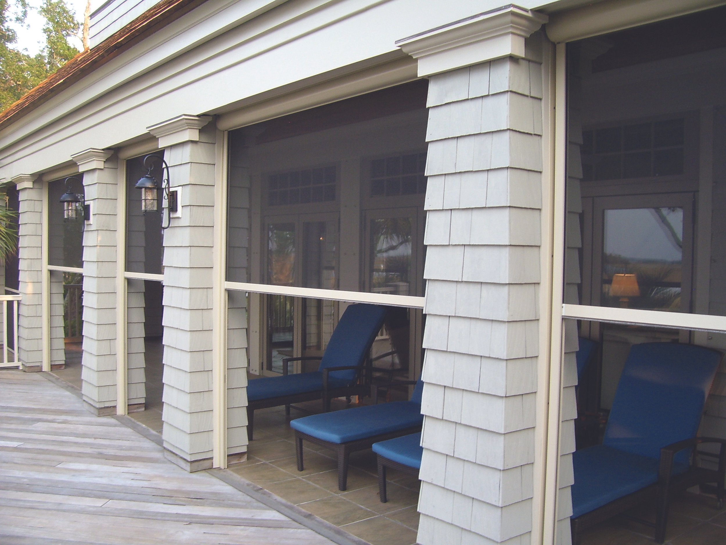 View of a house's screened porch with blue lounge chairs, decorative wall lights, and siding, overlooking a wooden deck and outdoor scenery.
