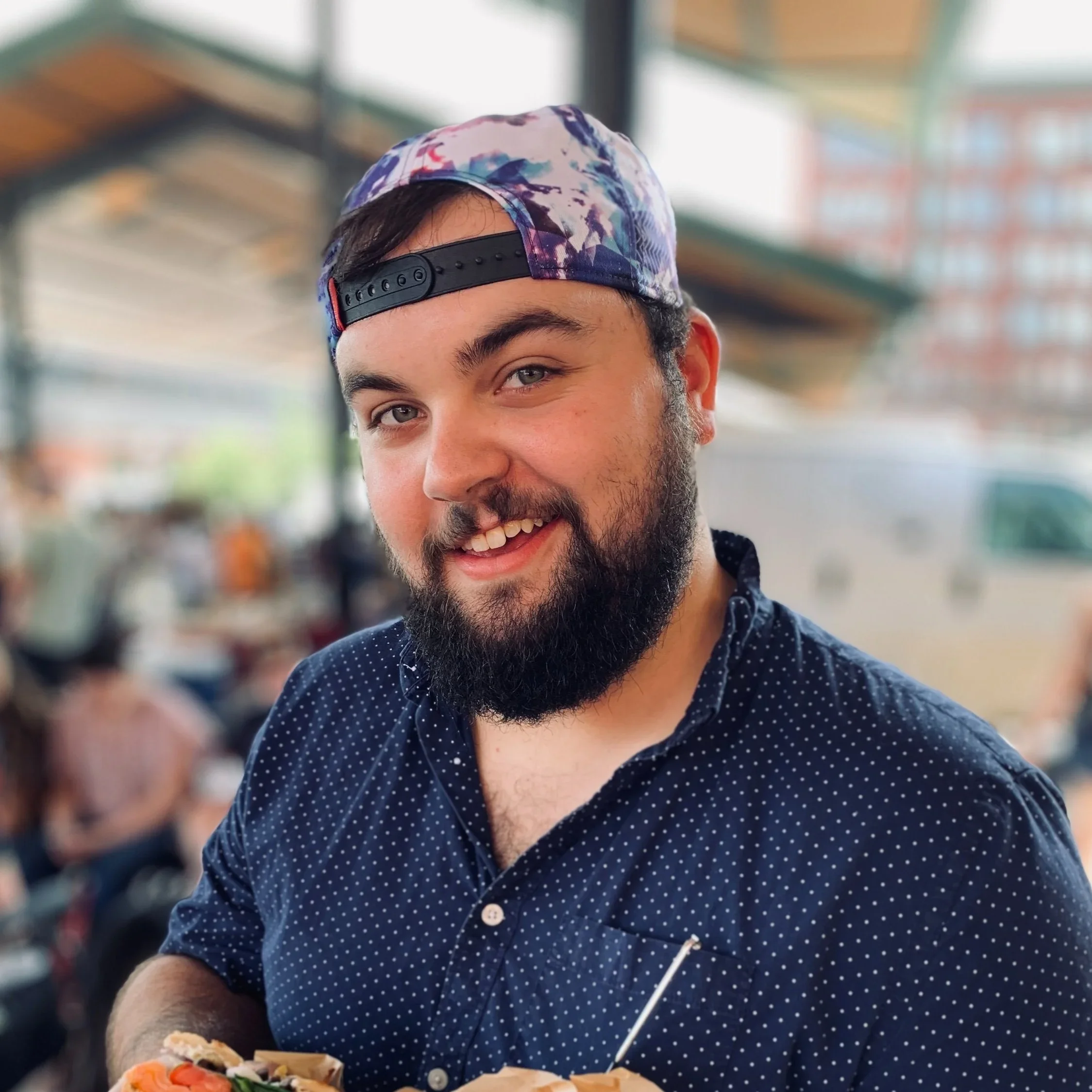 A young man with a beard, wearing a colorful cap backwards and a navy blue polka dot shirt, smiling outdoors with a blurred background of people and buildings.