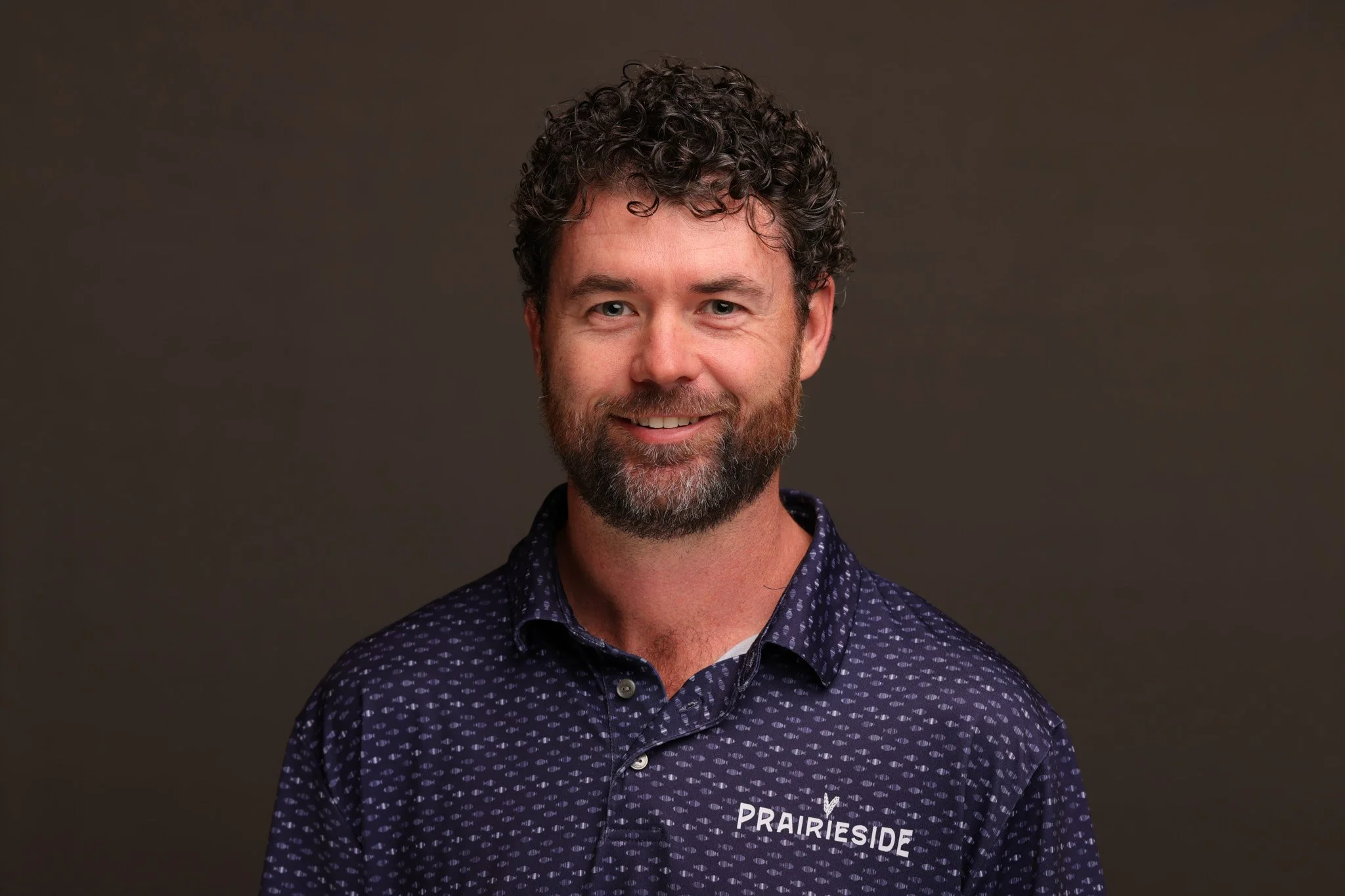 A man with curly dark hair, a beard, and a mustache, wearing a dark purple patterned polo shirt with the logo 'PRAIRIESIDE' on it, smiling against a plain dark background.