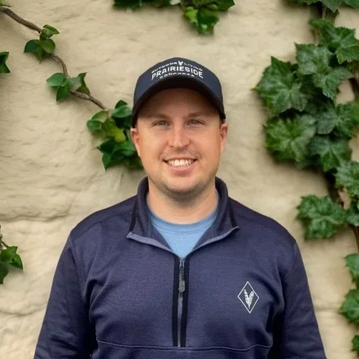 A smiling man wearing a dark blue jacket and a navy blue cap with 'PrairieSide' written on it, standing in front of a beige brick wall covered with green ivy.