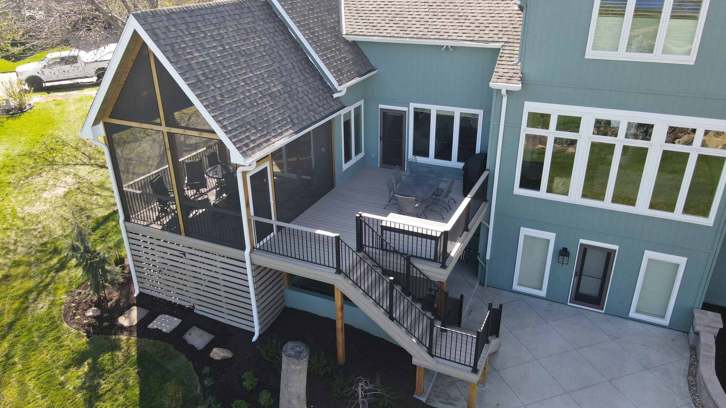 Aerial view of a house featuring a screened porch with outdoor furniture, a wooden deck, and a staircase leading down to a landscaped yard with grass and small plants.