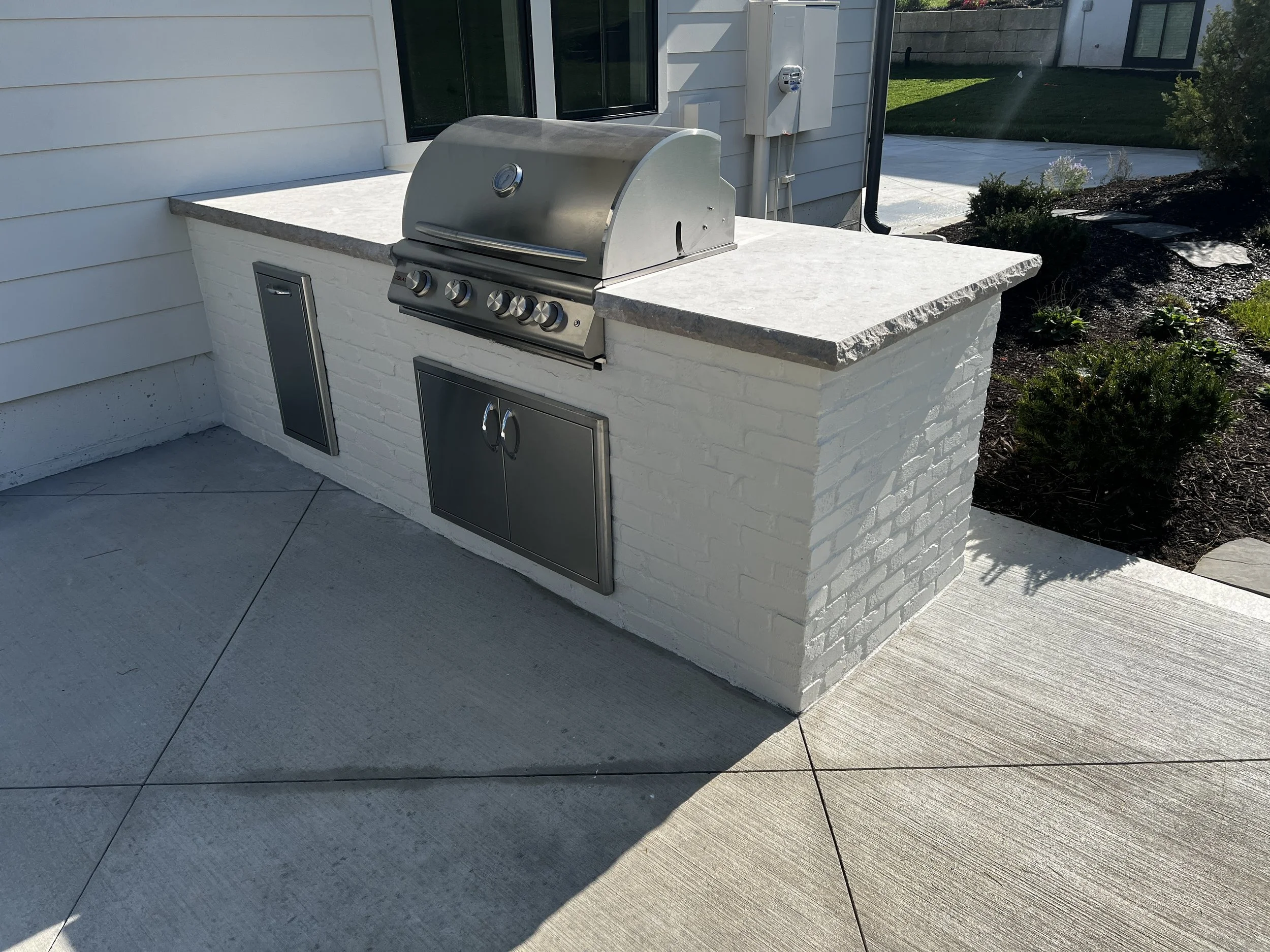Outdoor kitchen with stainless steel grill on a white brick counter, built into a white exterior wall, with cabinets and a concrete countertop, on a concrete patio.