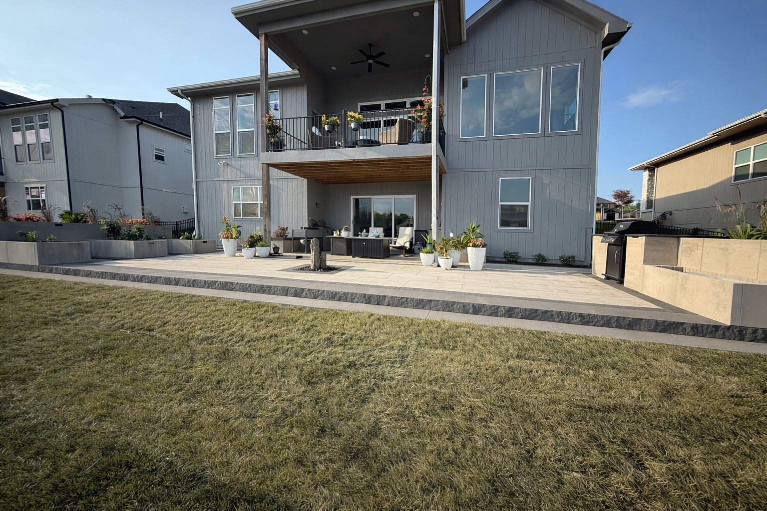 Rear view of a two-story house with a large patio, potted plants, outdoor seating, a barbecue grill, and neighboring houses in the background under a blue sky.