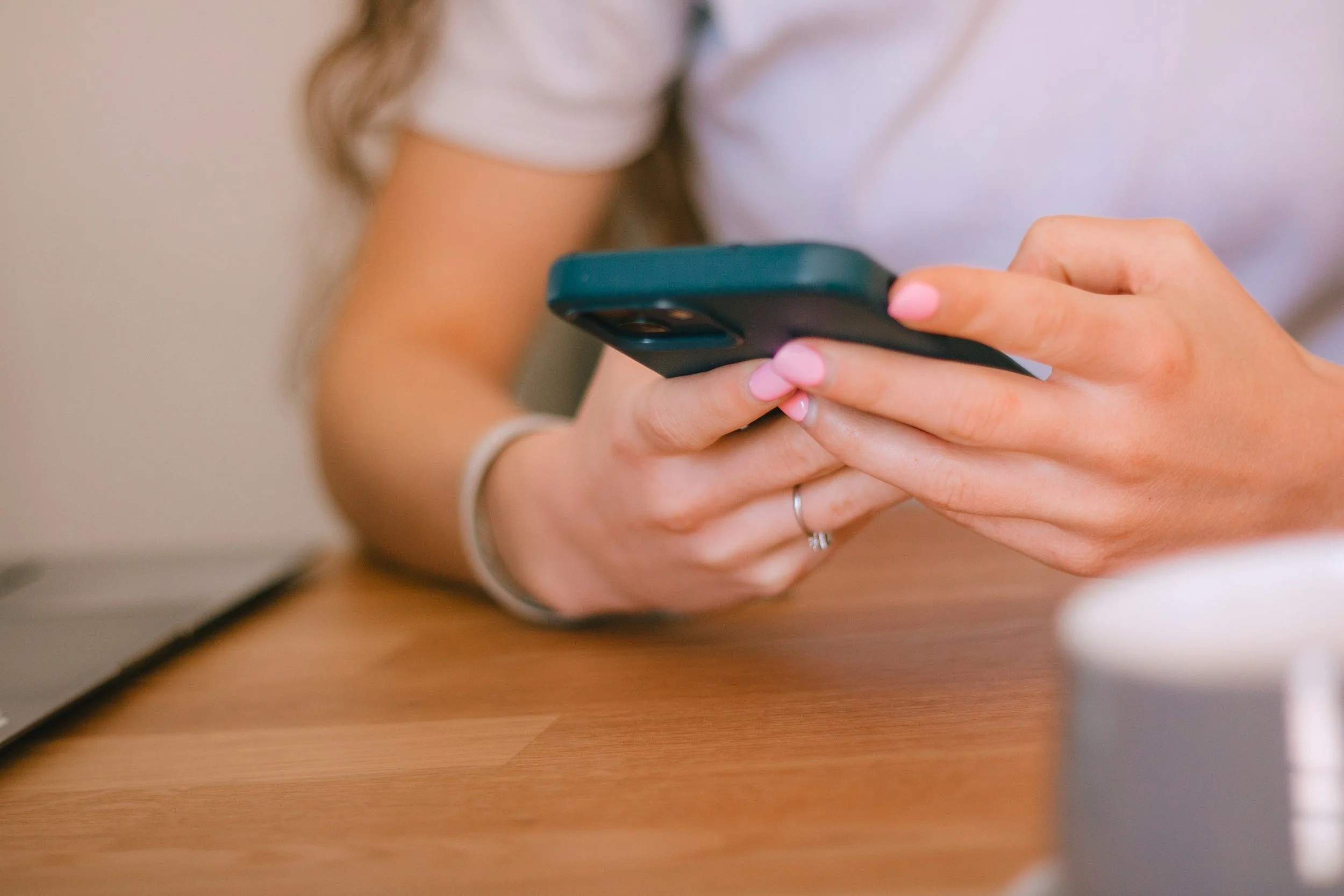 A woman with pink nail polish is holding and looking at a smartphone over a wooden table, with a laptop and a white cup in the background.