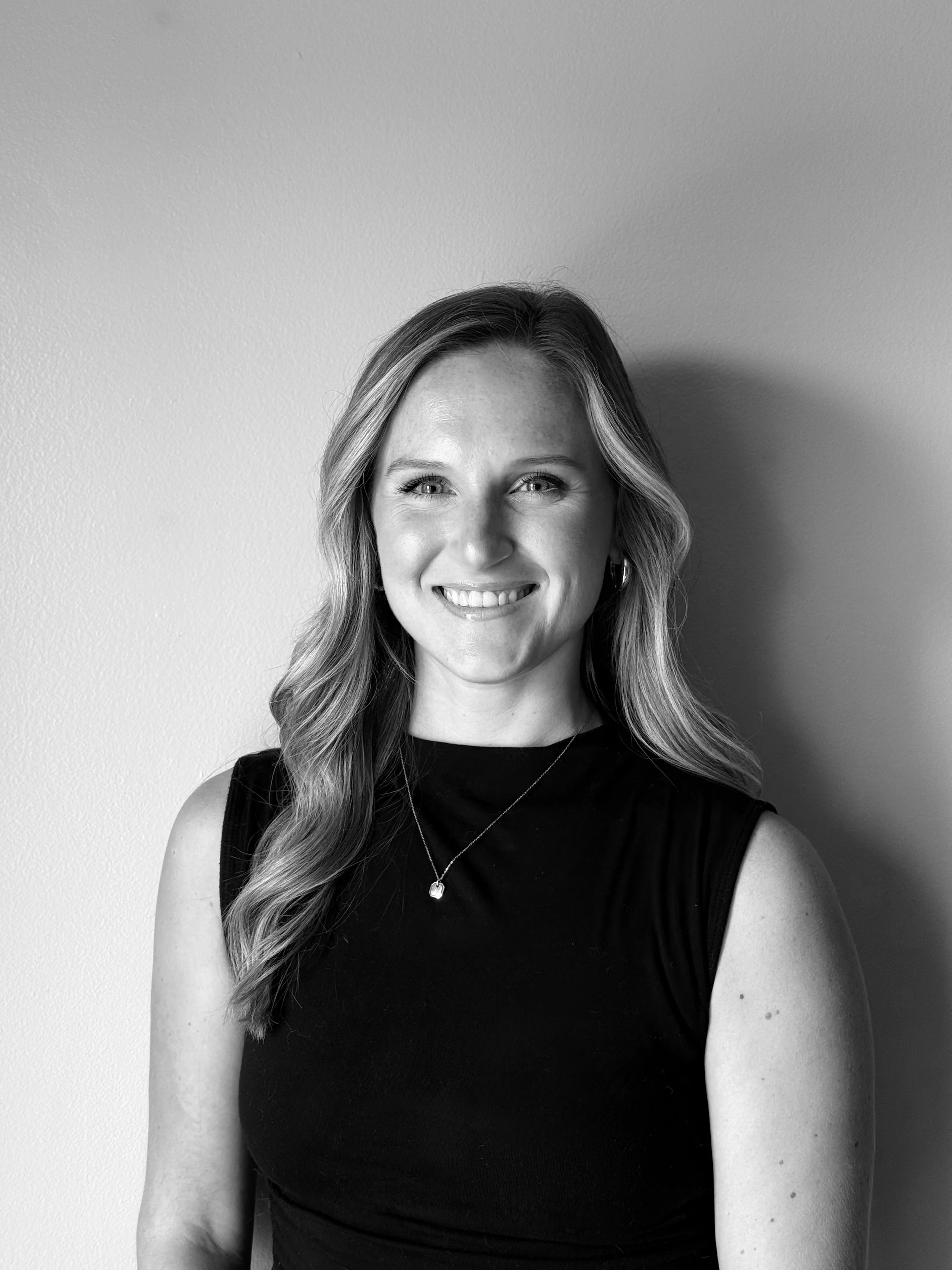 Black and white portrait of a smiling woman with shoulder-length wavy hair, wearing a sleeveless black top and a necklace, standing against a plain wall.