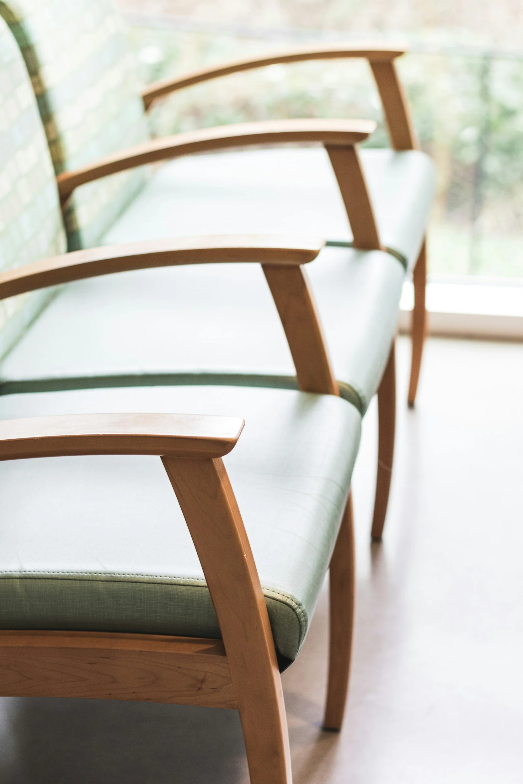 A row of three wooden chairs with light green cushions, positioned next to a window with a brick wall on the left.