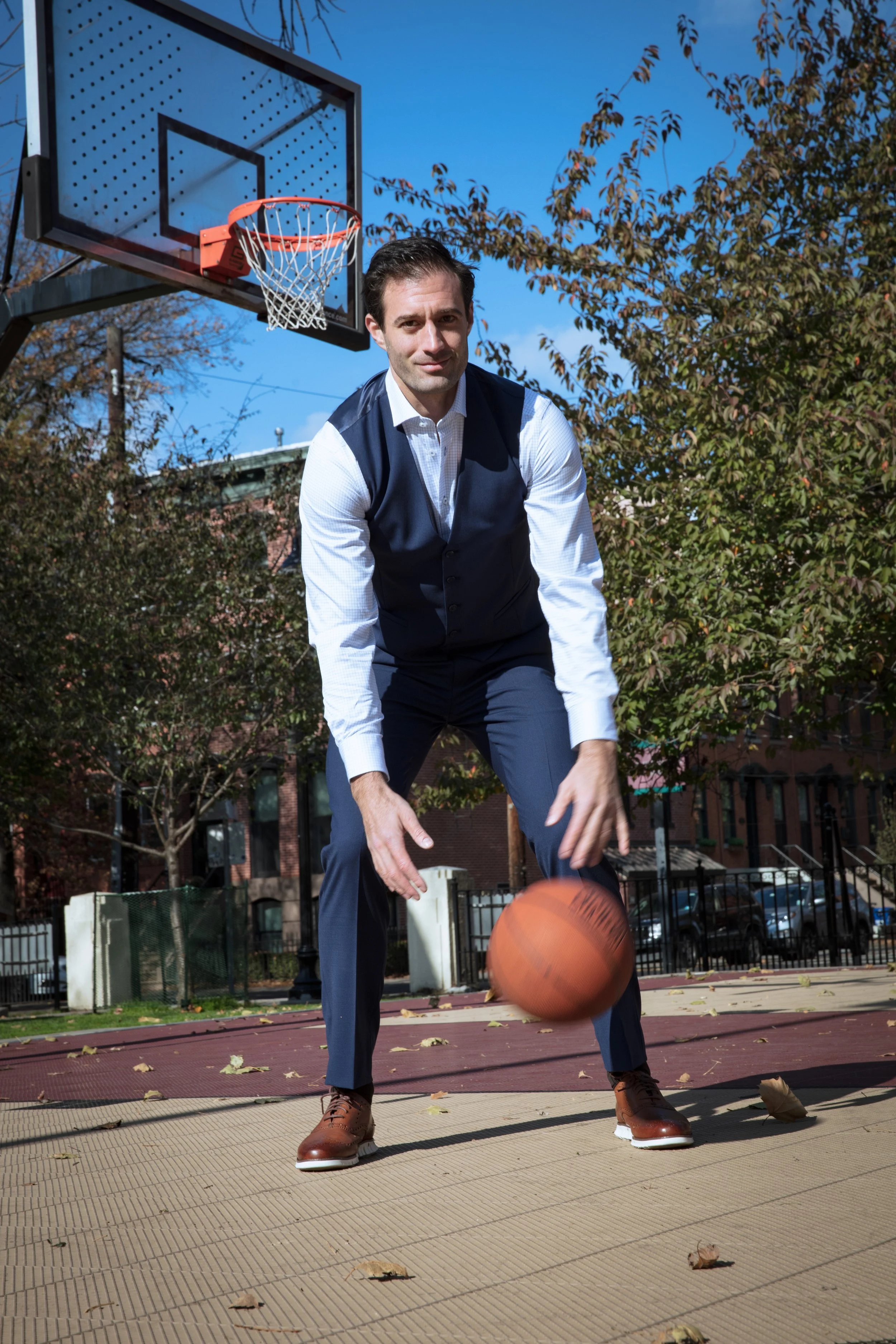 A man in business attire playing basketball on an outdoor court with trees and buildings in the background.