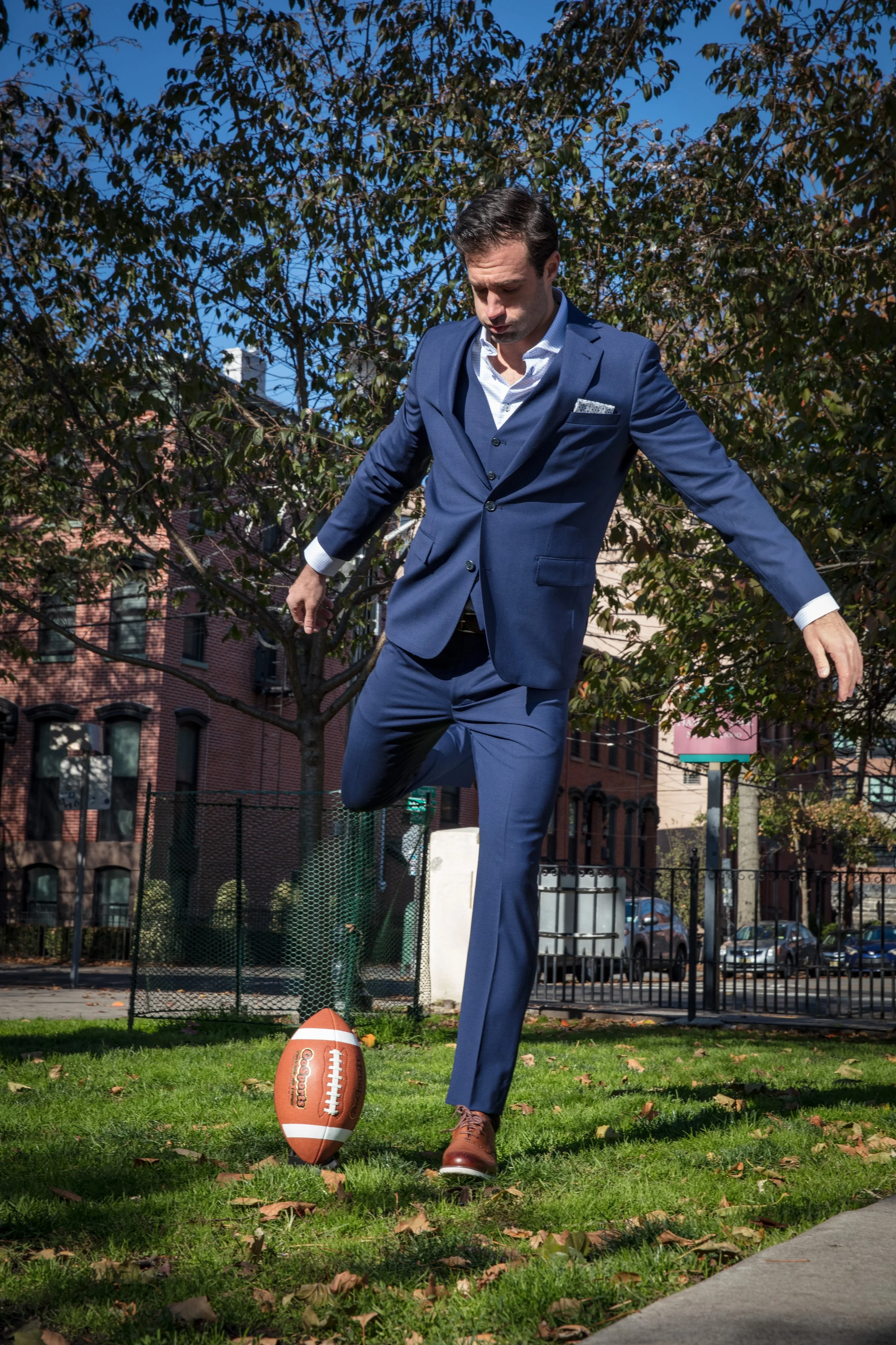 A man in a blue suit kicking a football in a park on a sunny day.
