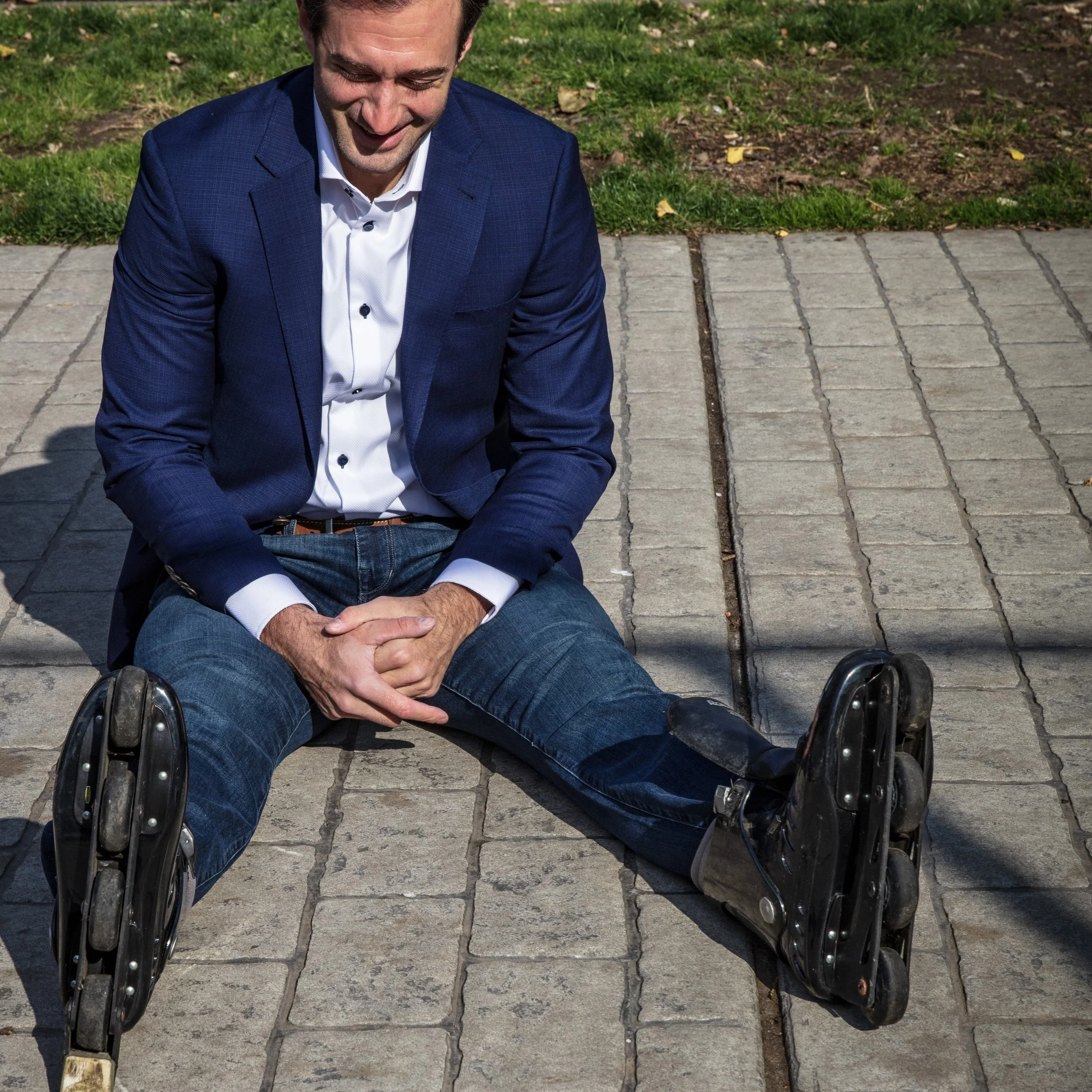 A man in a blue blazer and jeans sitting on the ground after falling, with his legs extended and rollerblades on.