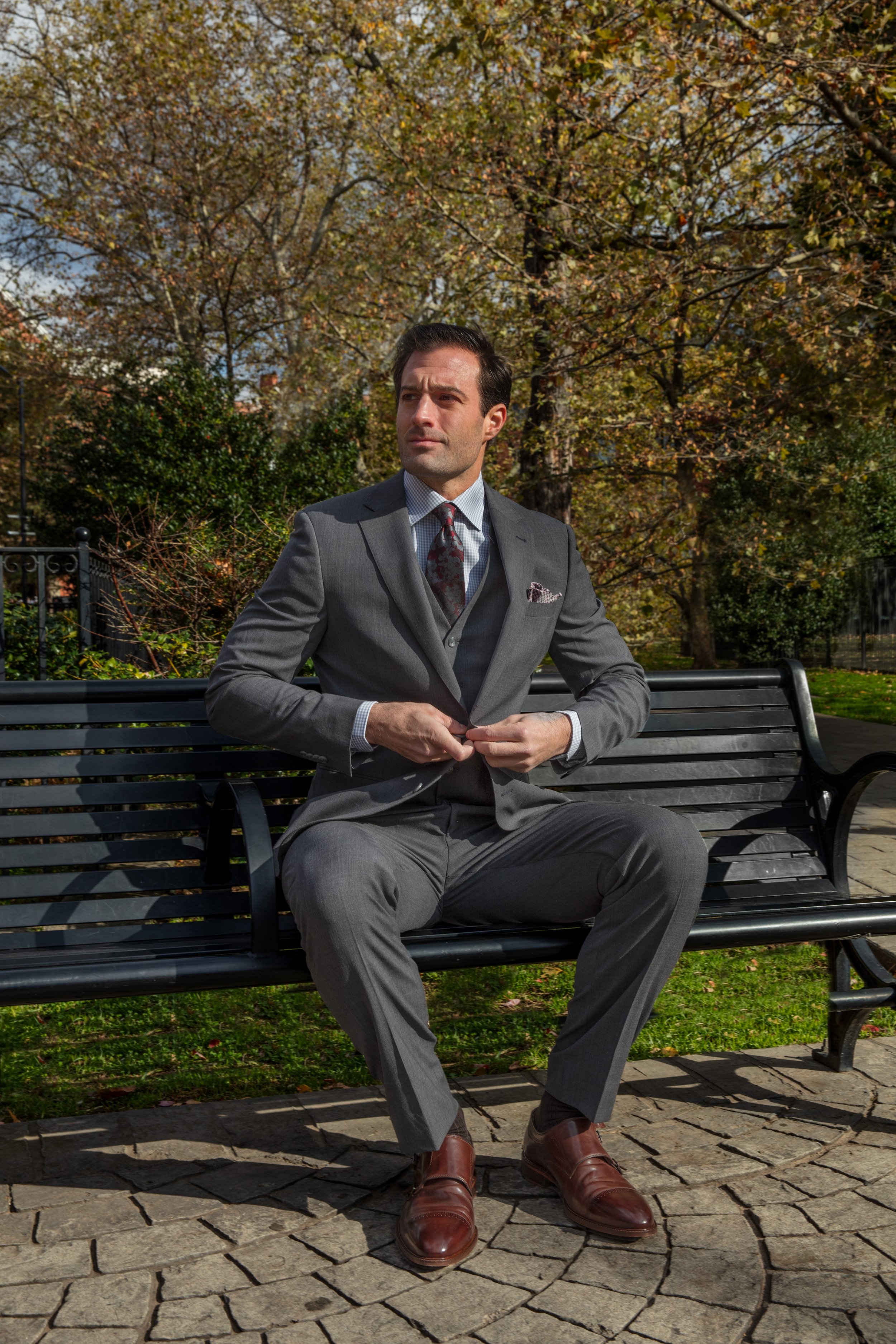 A man in a gray suit sitting on a park bench adjusting his jacket, with trees and autumn foliage in the background.