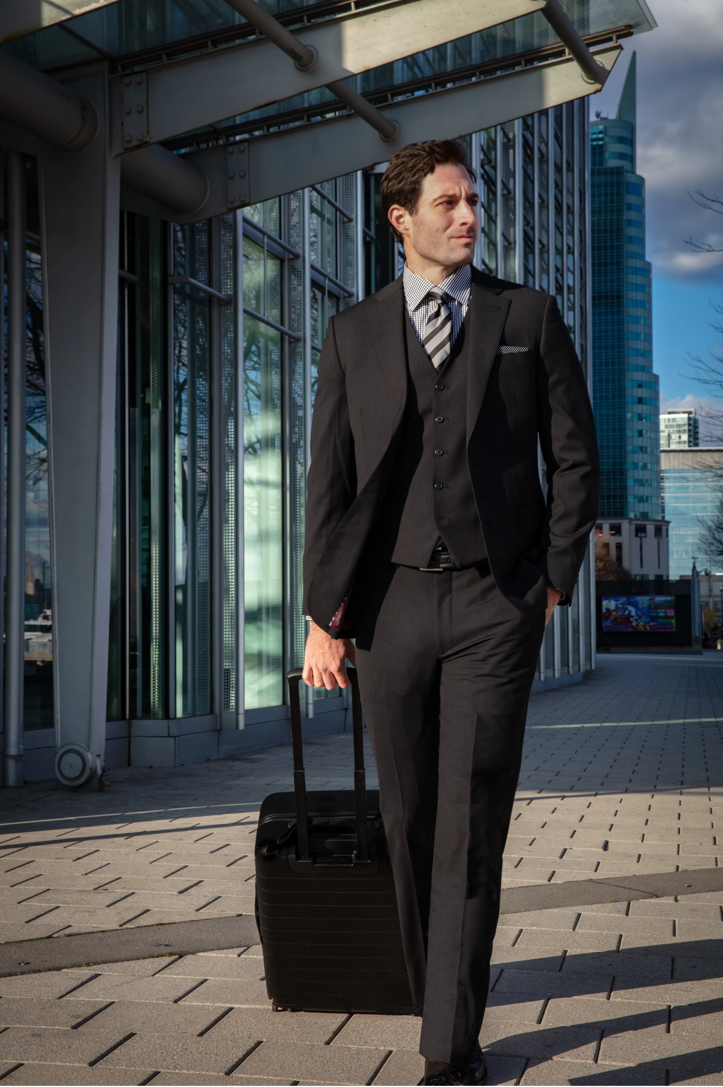 A man in a black suit walks outside a modern office building, pulling a black rolling suitcase. The city skyline is visible in the background.