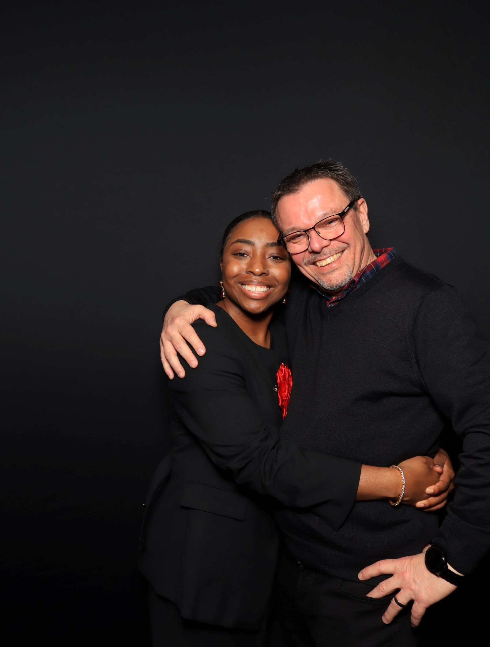 A man and woman hugging and smiling against a black background.