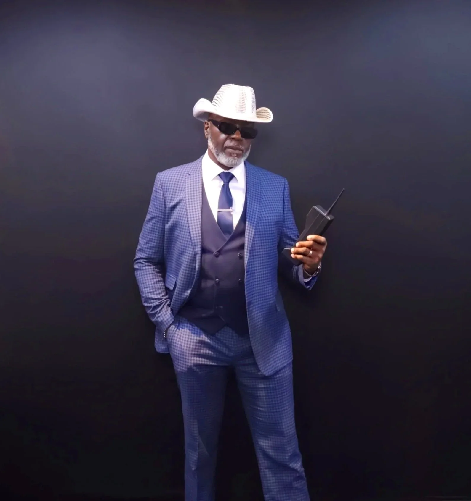 A man wearing a blue checkered suit, a white shirt, a navy vest, a matching tie, and sunglasses, standing against a black background. He has a white wide-brimmed hat with a decorative band and is holding a walkie-talkie in his right hand.