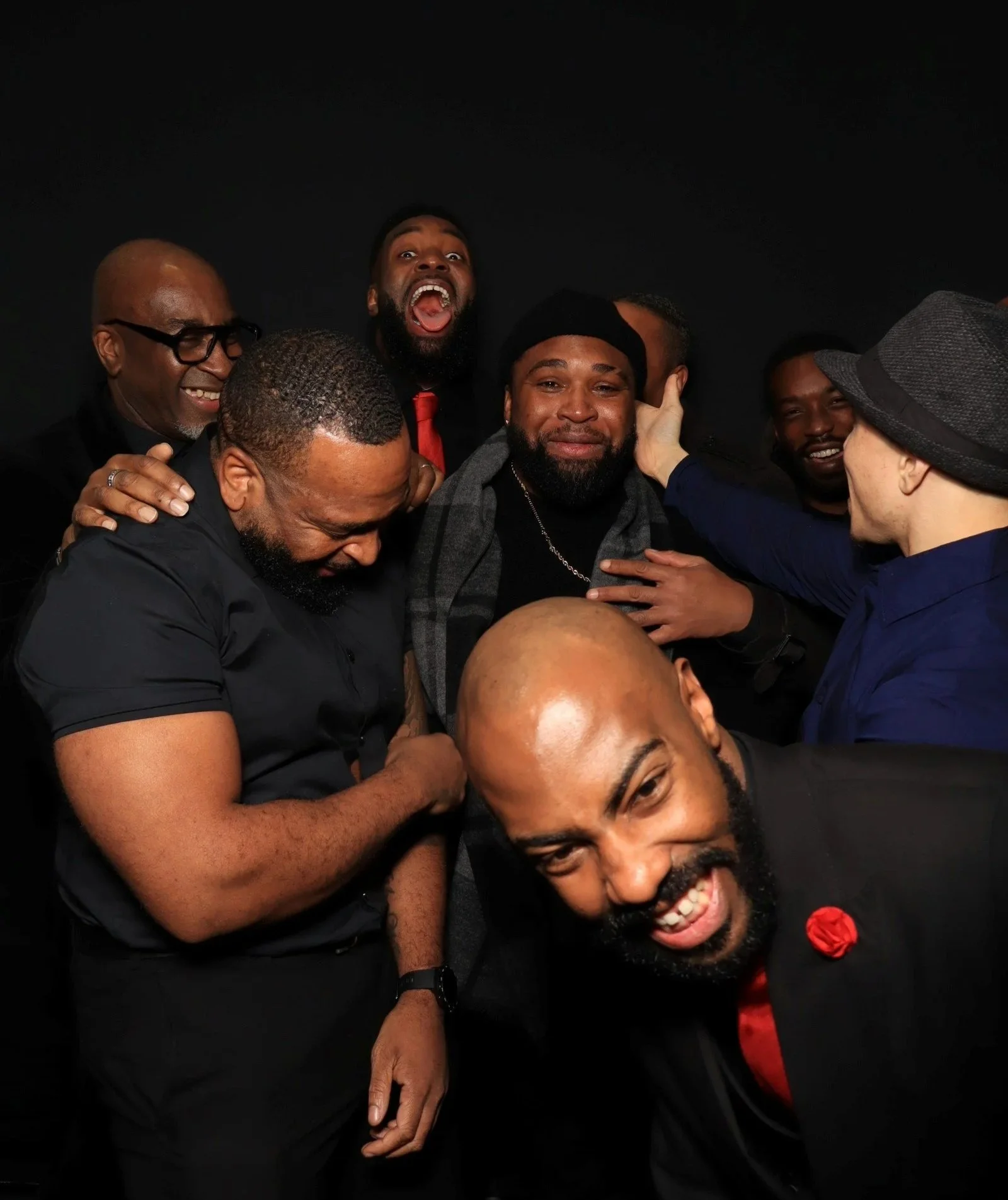 Group of seven men smiling and laughing together in a close, lively pose against a black background.