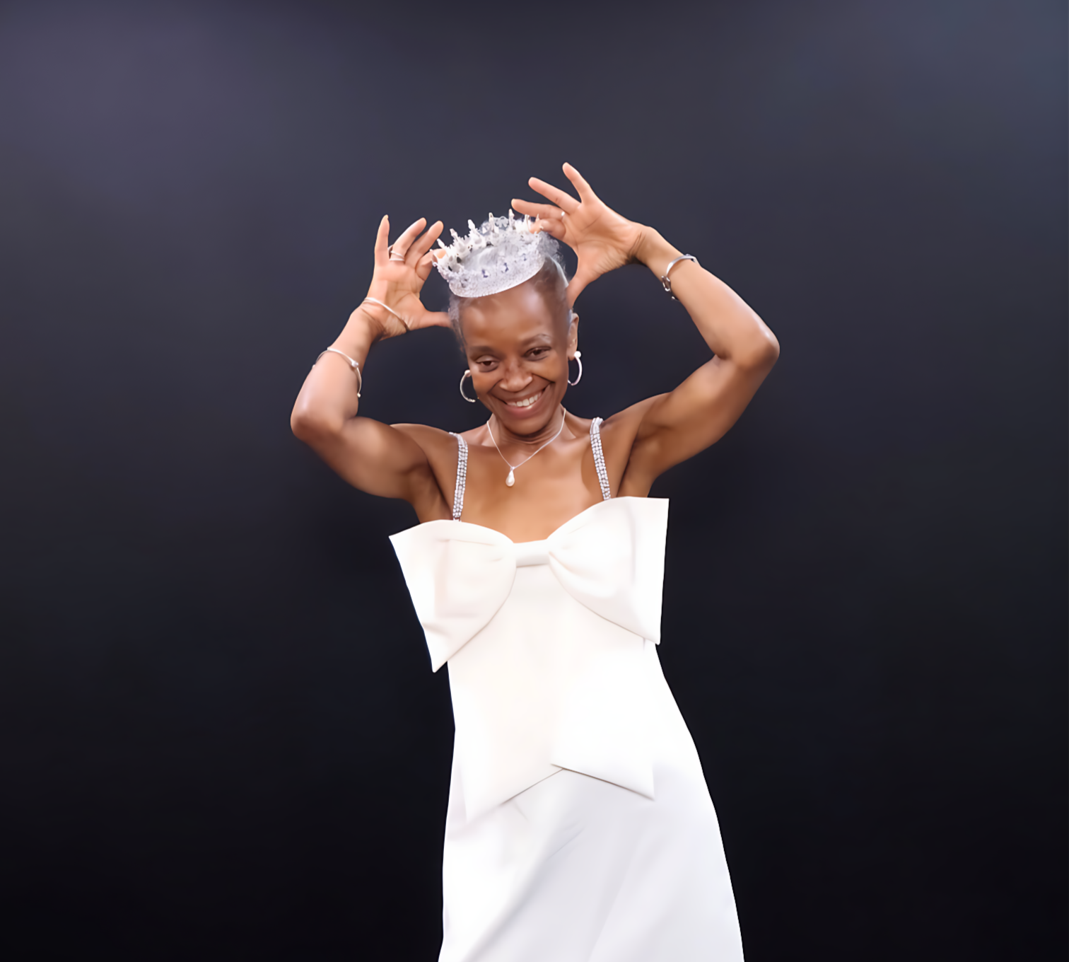A smiling woman in a white dress with bow details, wearing jewelry, places a tiara on her head against a black background.