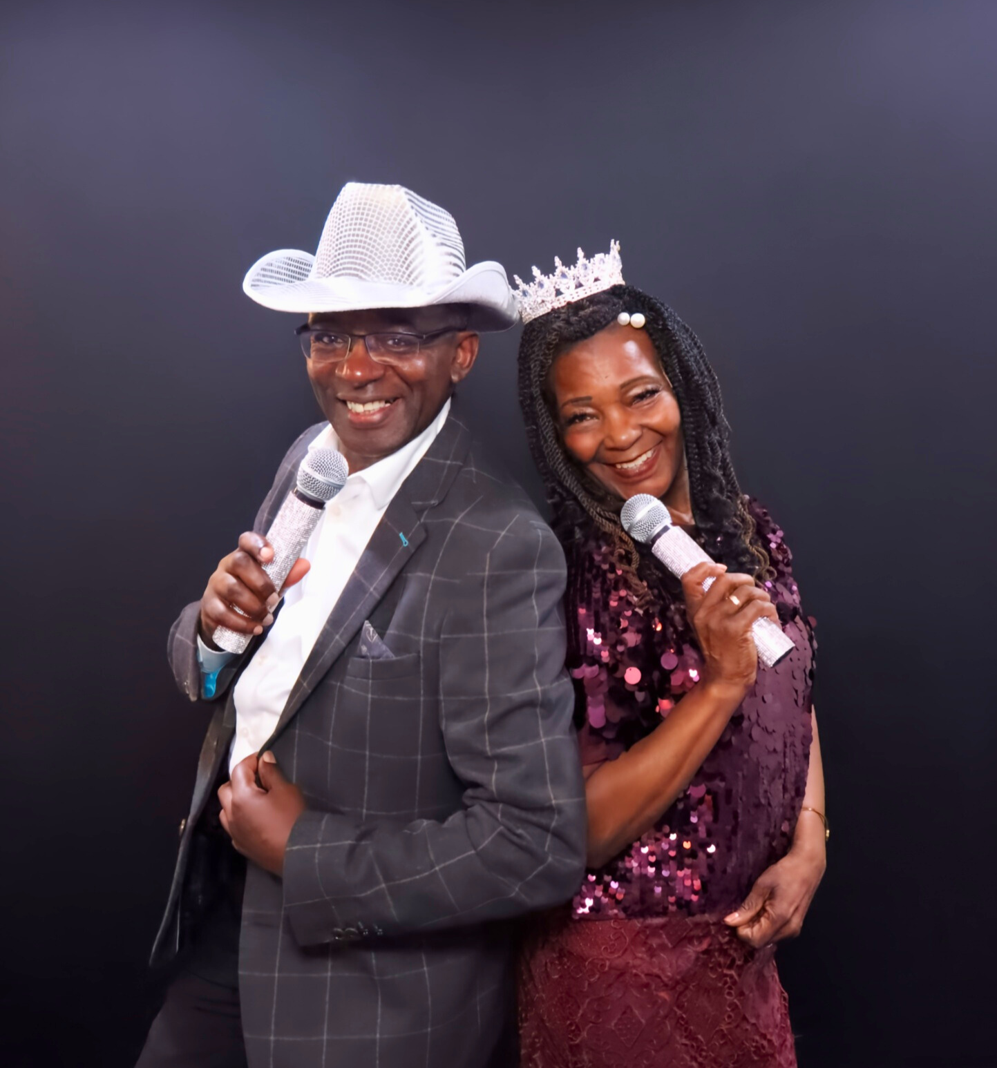A man and woman standing back to back smiling, each holding a microphone, dressed festively with the man in a cowboy hat and the woman wearing a crown and a sequined top.