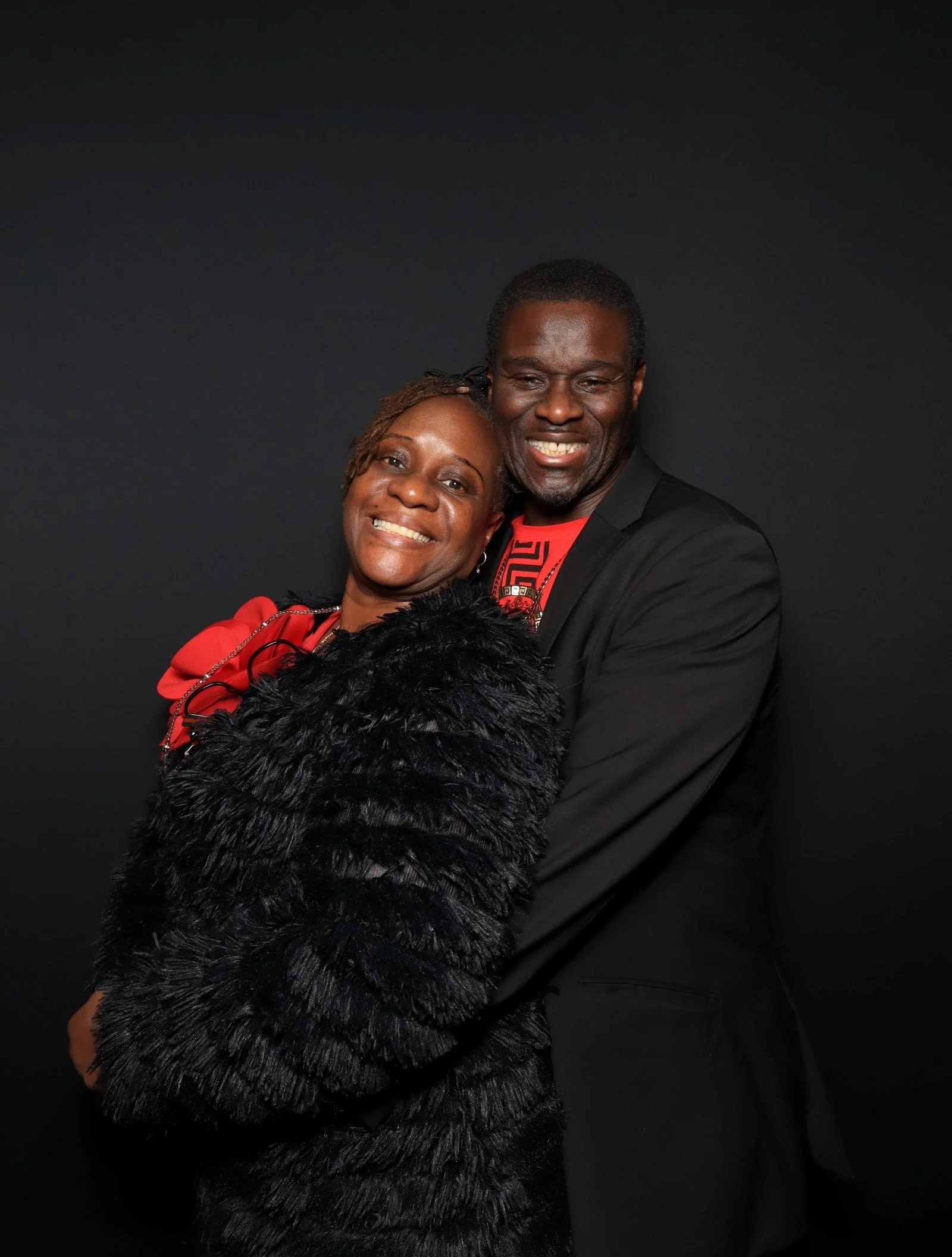 A smiling couple embracing against a black background, dressed in formal attire, with the woman wearing a red dress and black fur stole, and the man wearing a black suit with a red shirt.