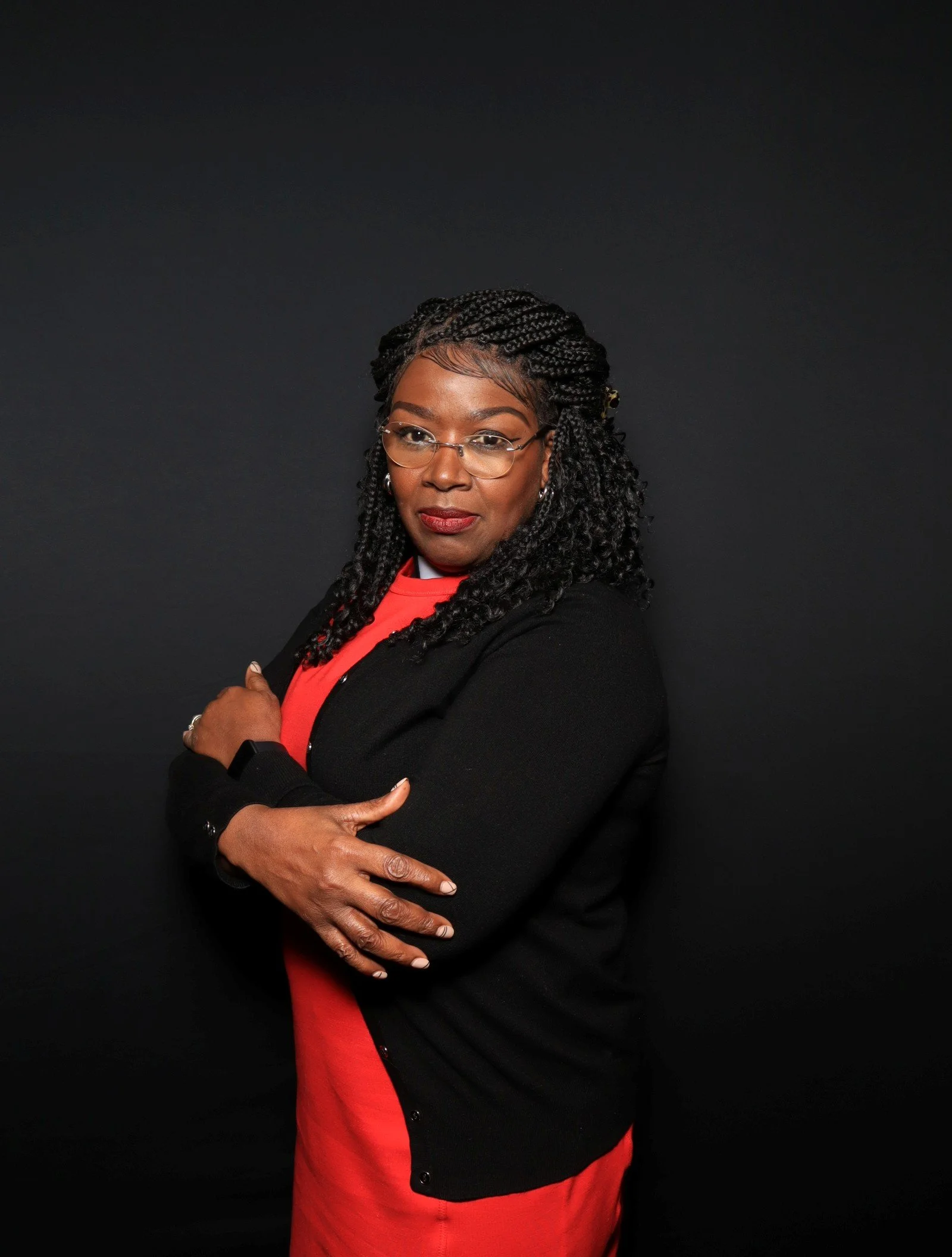 A confident African American woman with braided hair and glasses standing with arms crossed against a dark background, wearing a red dress and black blazer.