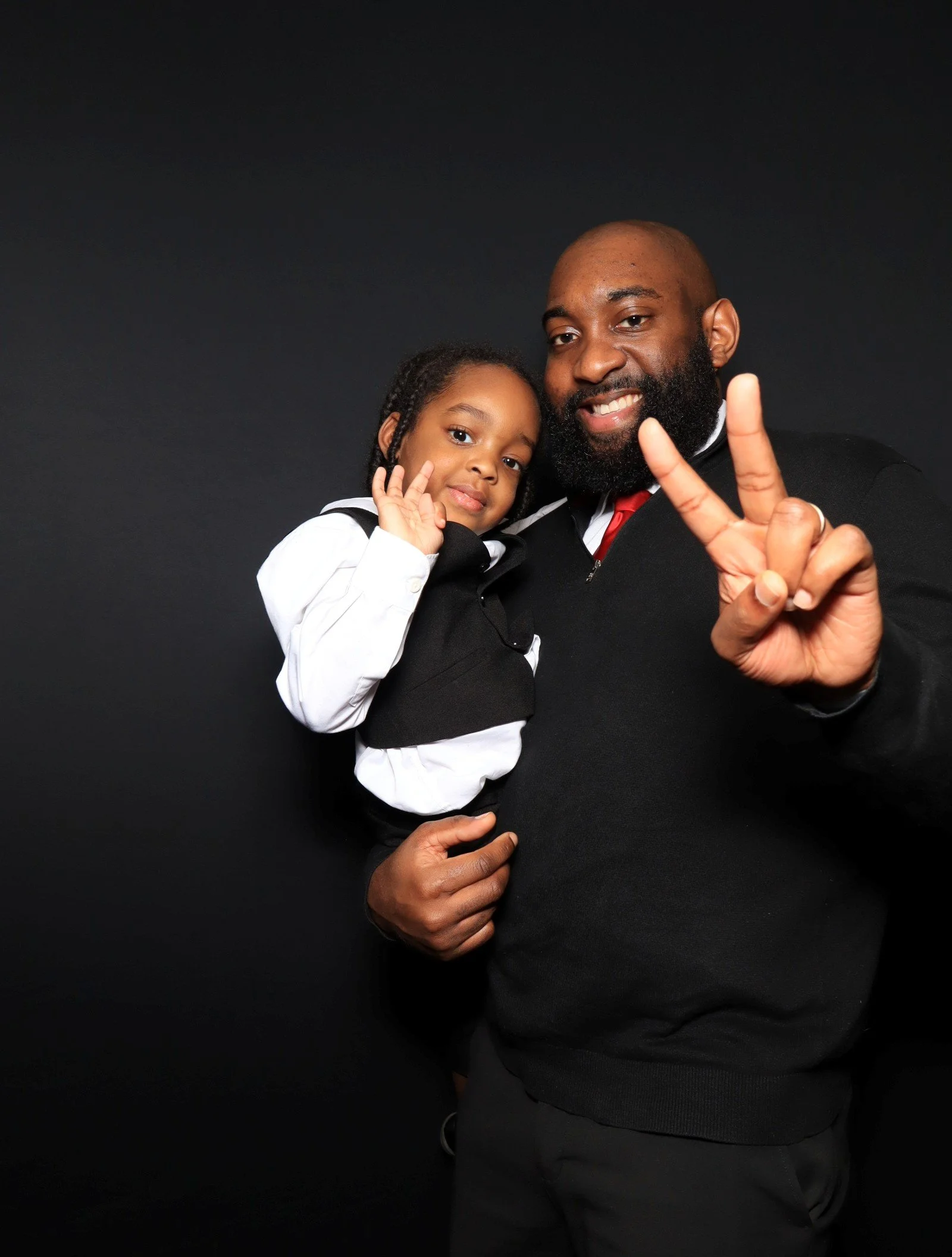 A man with a beard holding a young girl, both smiling and making a peace sign with his right hand, against a black background.