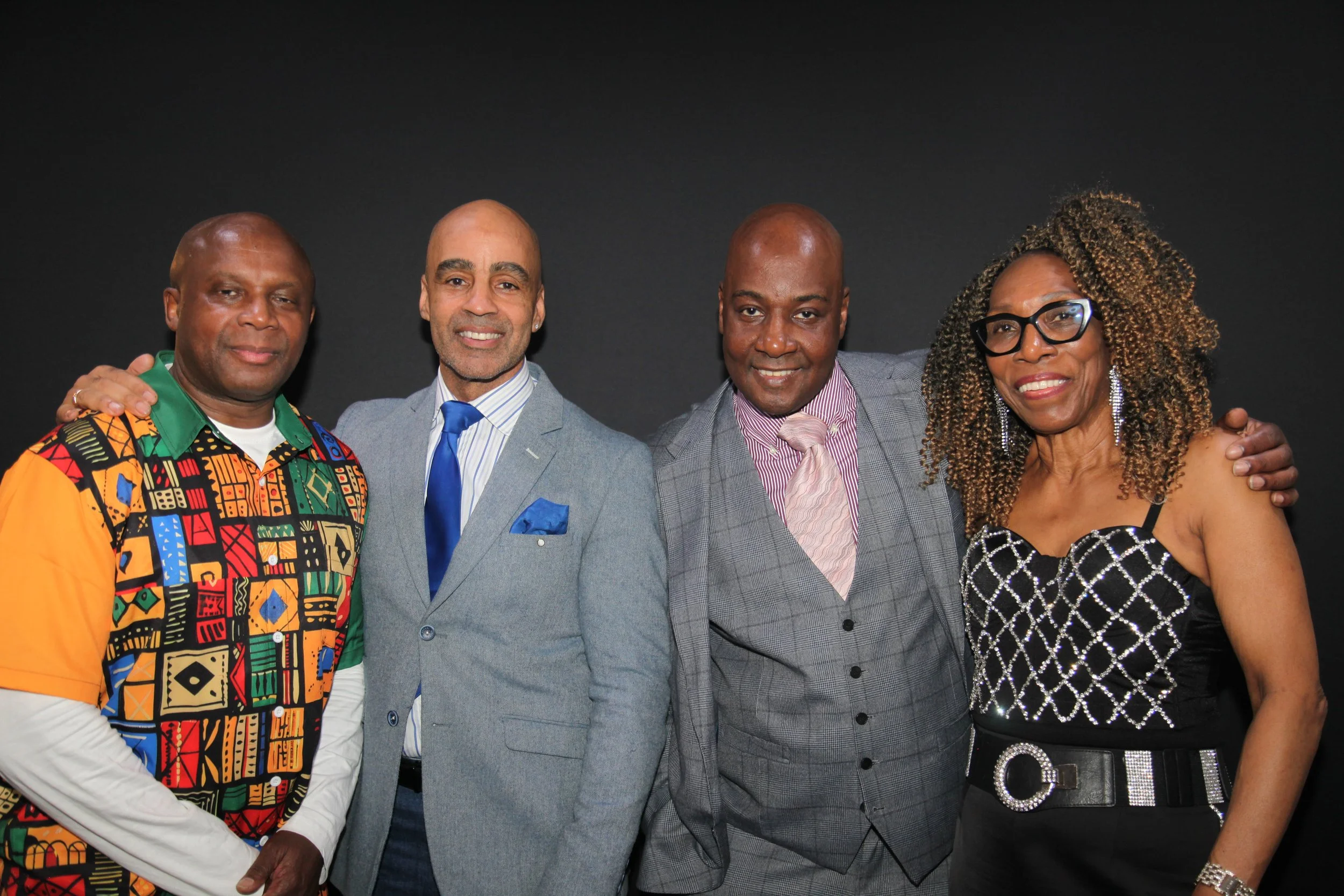 Four adults standing close together, smiling, against a black background. They are dressed in colorful and formal attire.