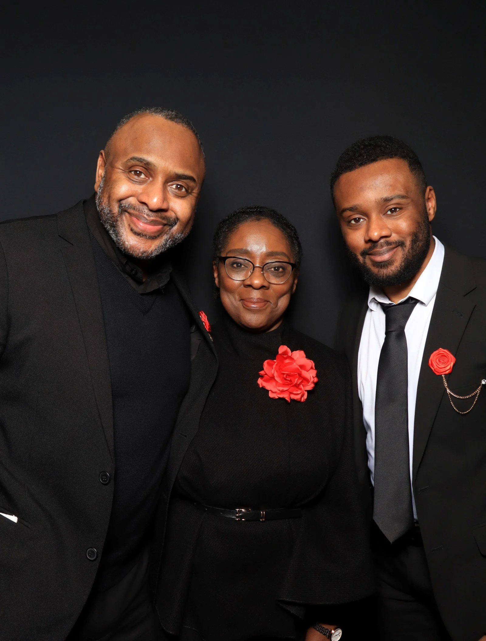Three people dressed in black formal attire, standing against a dark background, two men and one woman in the middle, all wearing red floral pins.