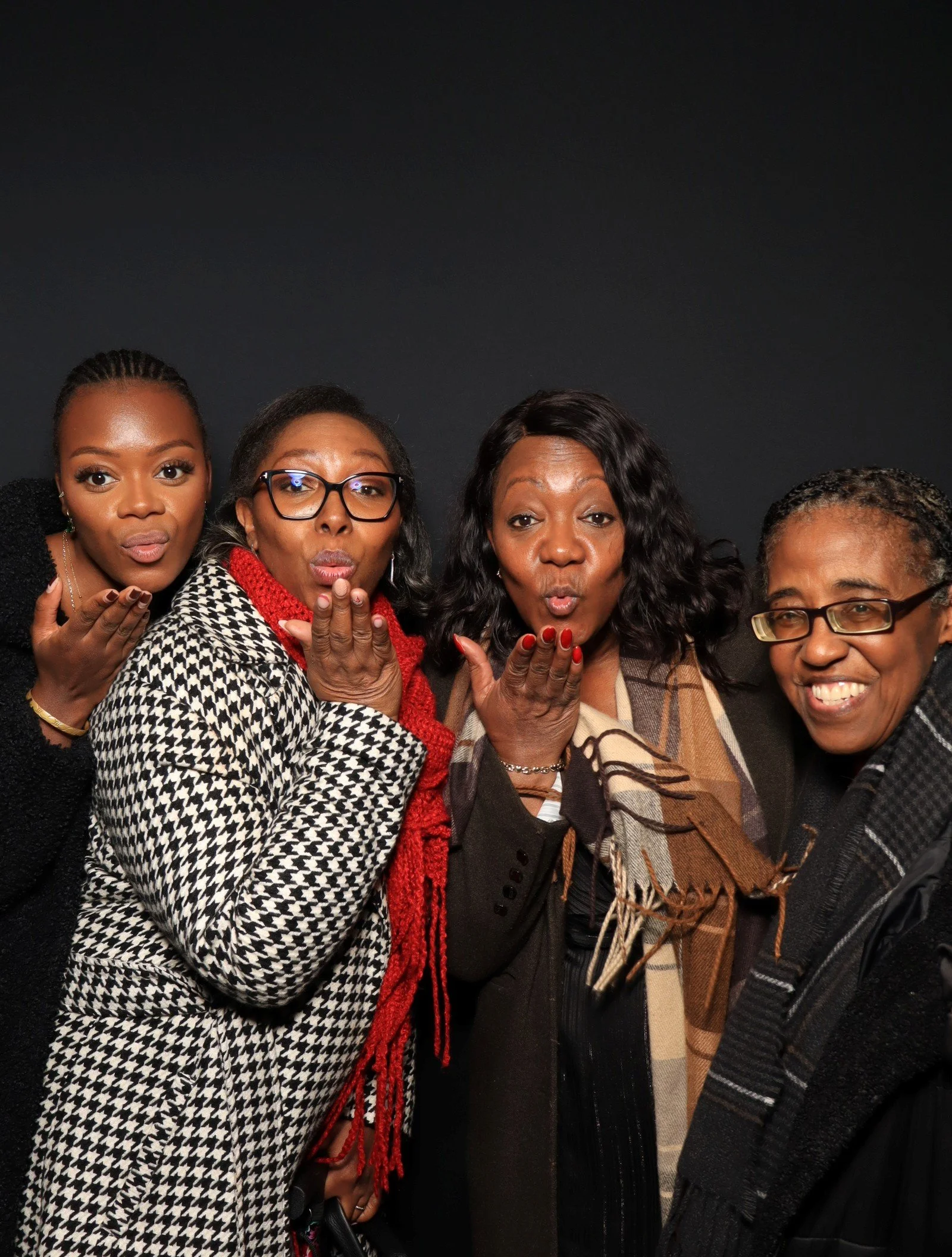 Four women blowing kisses at the camera against a black background.