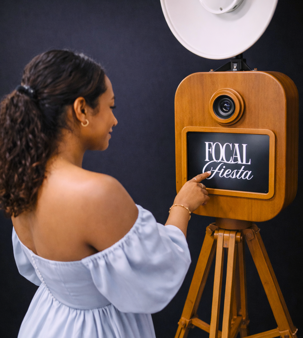 A woman with curly hair in a white off-shoulder dress interacts with a vintage wooden photo booth camera displaying the words "Focal Fiesta" on a screen.