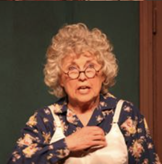 An elderly woman with curly gray hair and glasses, wearing a floral shirt and white apron, standing in front of a green background.