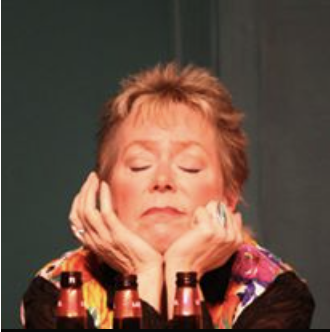 A woman with short, reddish hair resting her chin on her hands among small bottles of nail polish, with a calm or contemplative expression.