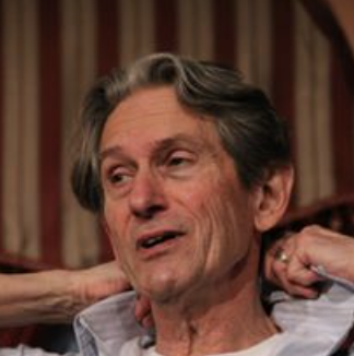 An older man with gray hair speaking or in conversation, wearing a light-colored shirt, with a striped curtain background.