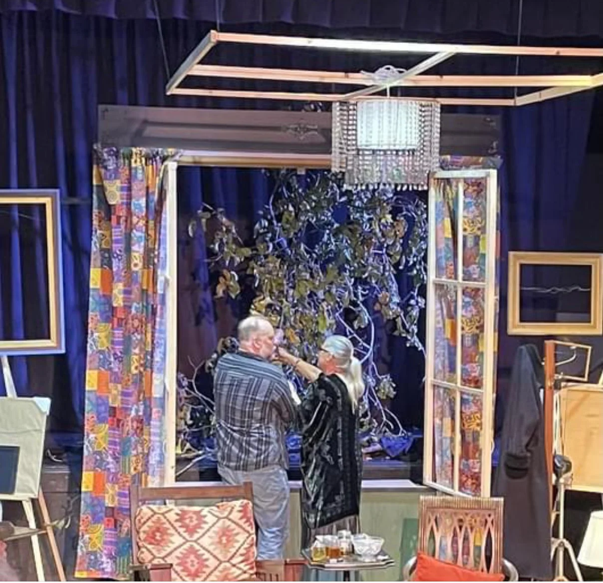 Two elderly women talking in front of a stage decorated with a tree, open windows, colorful curtains, picture frames, chairs, a table with drinks, and a chandelier.