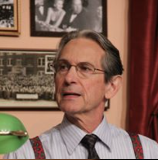 A man with glasses and dark hair wearing a striped shirt, black tie, and suspenders, standing in a room with framed photos and a red curtain behind him.
