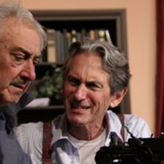 Two elderly men smiling and looking at each other indoors, with bookshelves in the background.