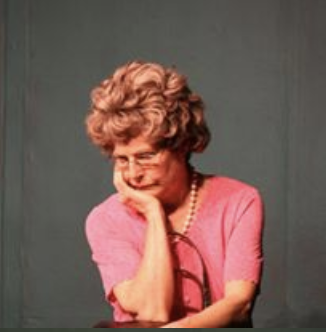 A woman with curly gray hair, wearing glasses and a pink shirt, resting her chin on her hand.