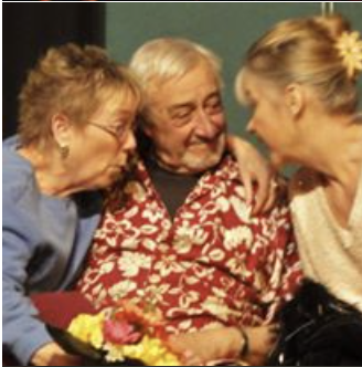 Three elderly people sitting close together and smiling, with two women on either side of a smiling man in the middle. The woman on the right is holding a bouquet of flowers.