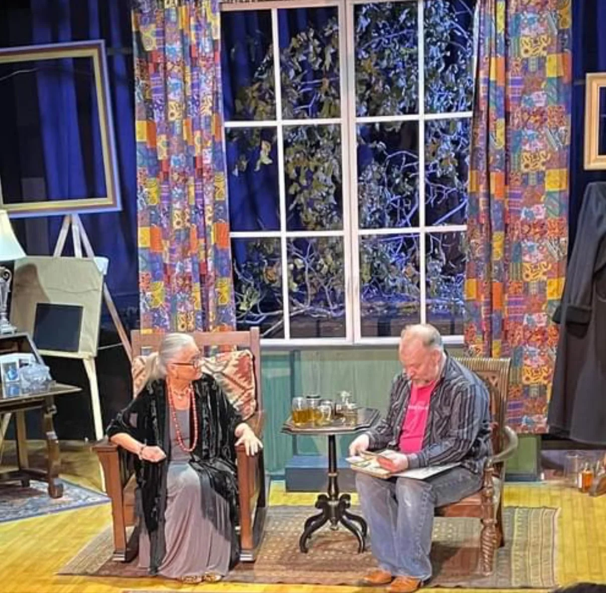 A room with two elderly people sitting in wooden chairs, having a conversation. The woman has long white hair, glasses, and is wearing a black patterned cardigan and gray dress. The man has short gray hair, glasses, and is dressed in a striped blazer