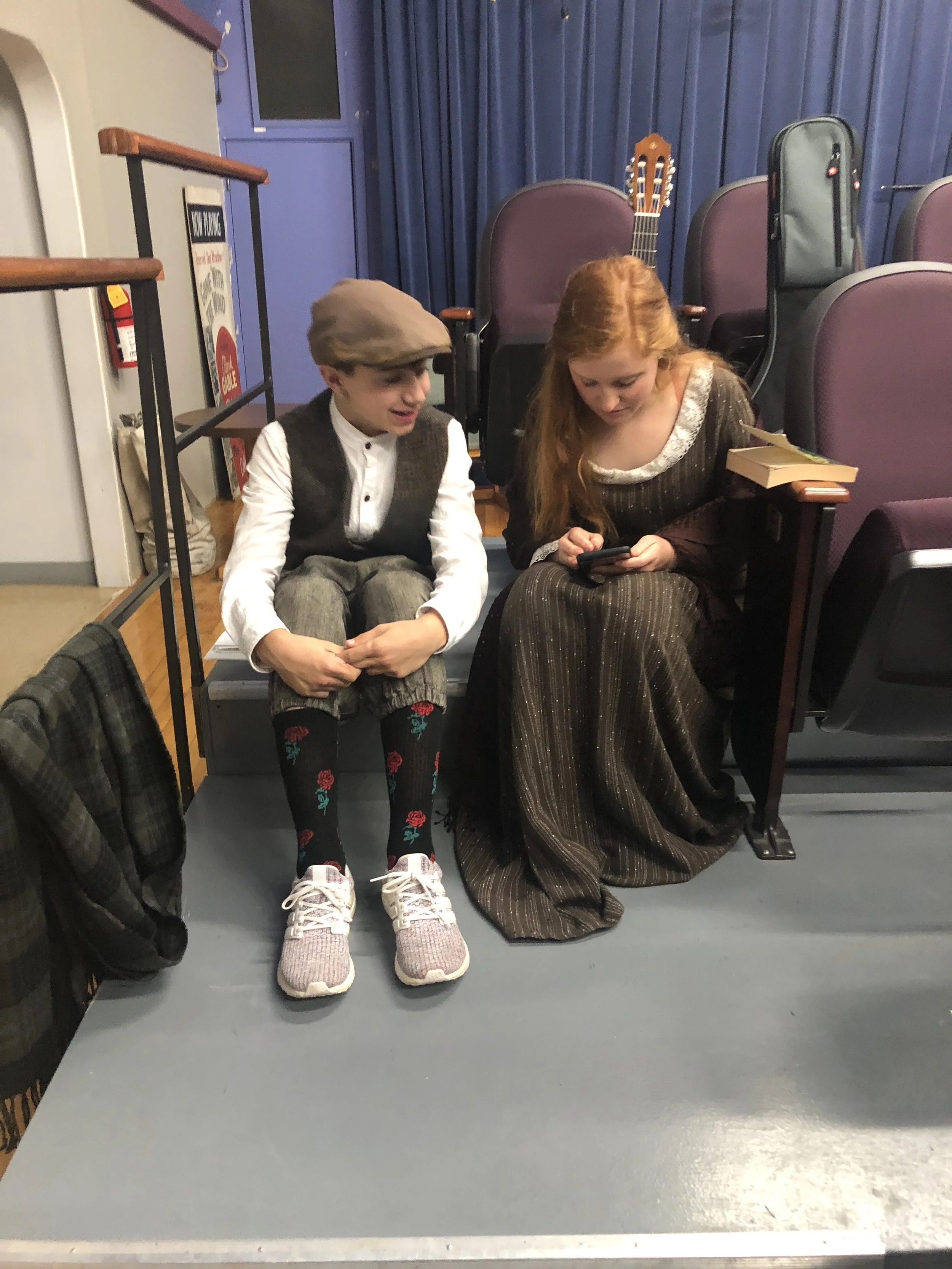 A young boy and girl sitting on a stage, looking at a phone together, with purple theater chairs and a guitar in the background.