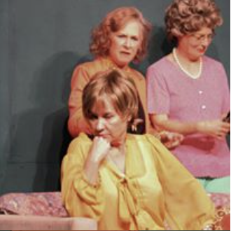 Three women sitting together, with one woman in a yellow blouse looking thoughtful, and the other two women in pink and orange tops smiling.