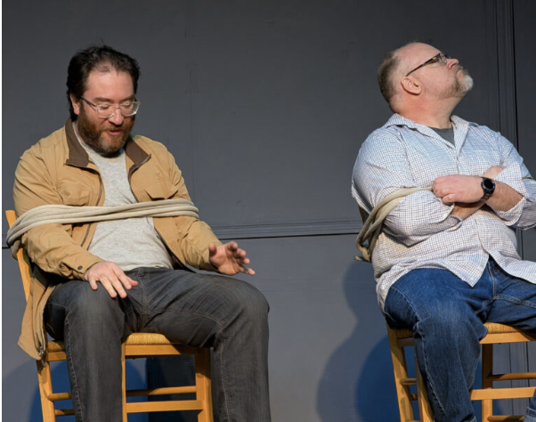 Two men sitting on chairs with their hands tied behind their backs and wrapped with ropes, appearing to be participating in a staged performance or activity.