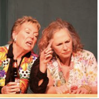 Two women sitting at a bar, one is talking animatedly while the other is listening thoughtfully, with drinks on the bar counter.