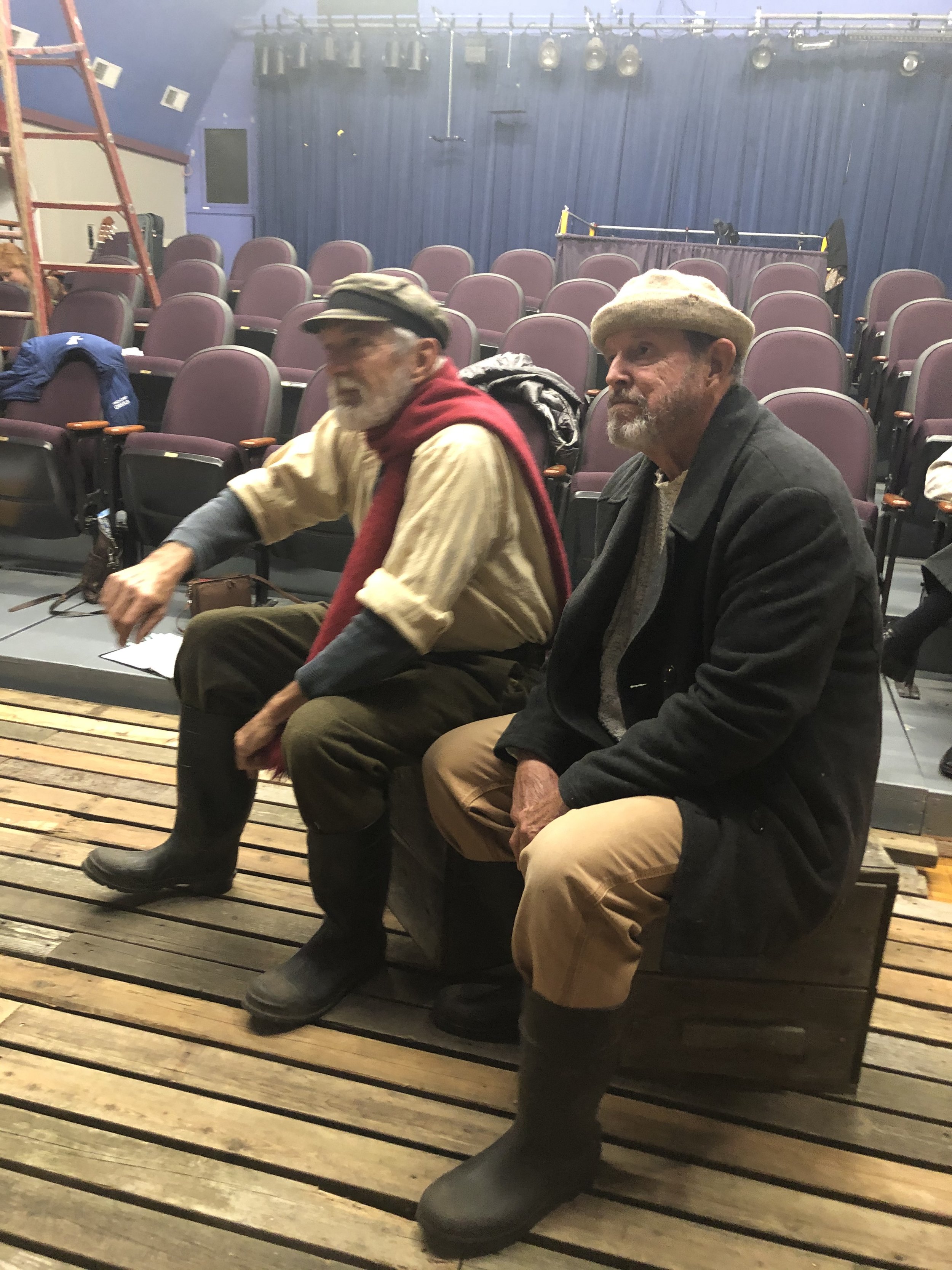 Two elderly men wearing vintage outdoor clothing are seated on a wooden bench with a stage and rows of empty purple seats in the background.