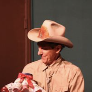 A man wearing a large cowboy hat and a beige shirt, holding a wrapped gift.