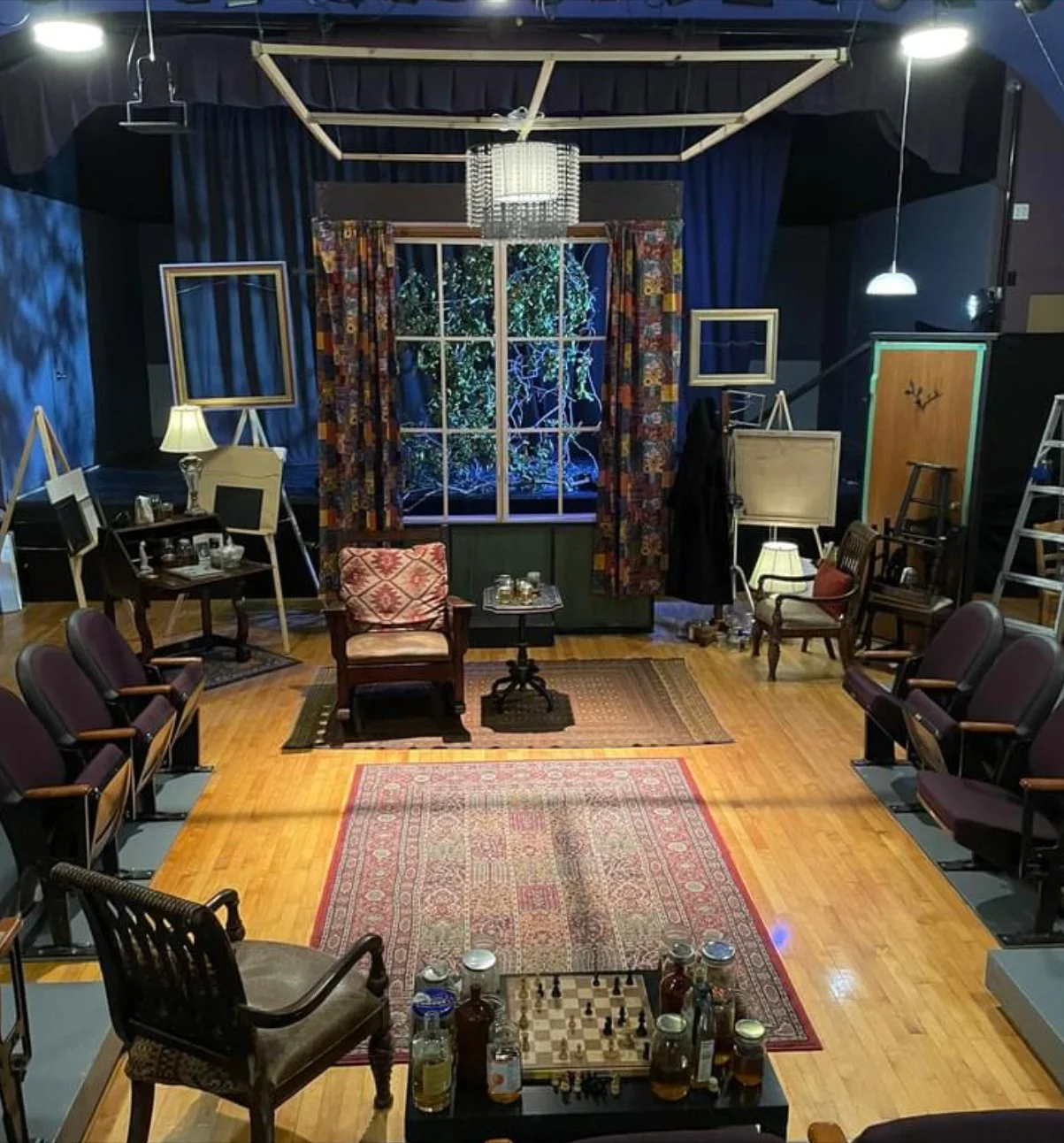 A theatrical or film stage setup that resembles a cozy living room with vintage furniture, lamps, a window with curtains, a Christmas tree behind the window, and various picture frames on the walls and floor.