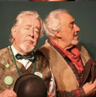 Two elderly men dressed in old-fashioned clothing, one with a bowtie and the other with a red scarf, sitting close together against a dark green background.